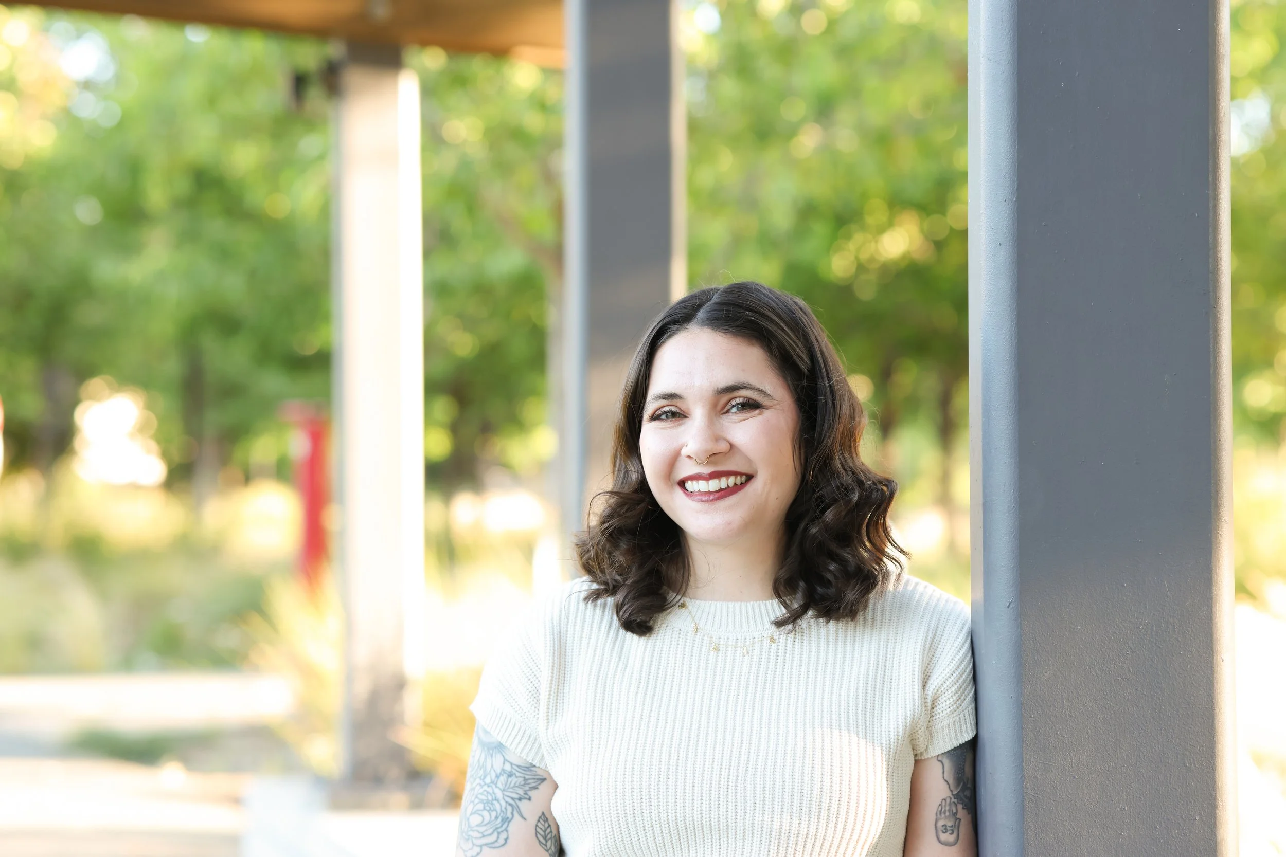 Portrait of a young woman with dark hair, smiling, wearing a white sweater, standing outdoors near a gray pillar in a park with green trees in the background.