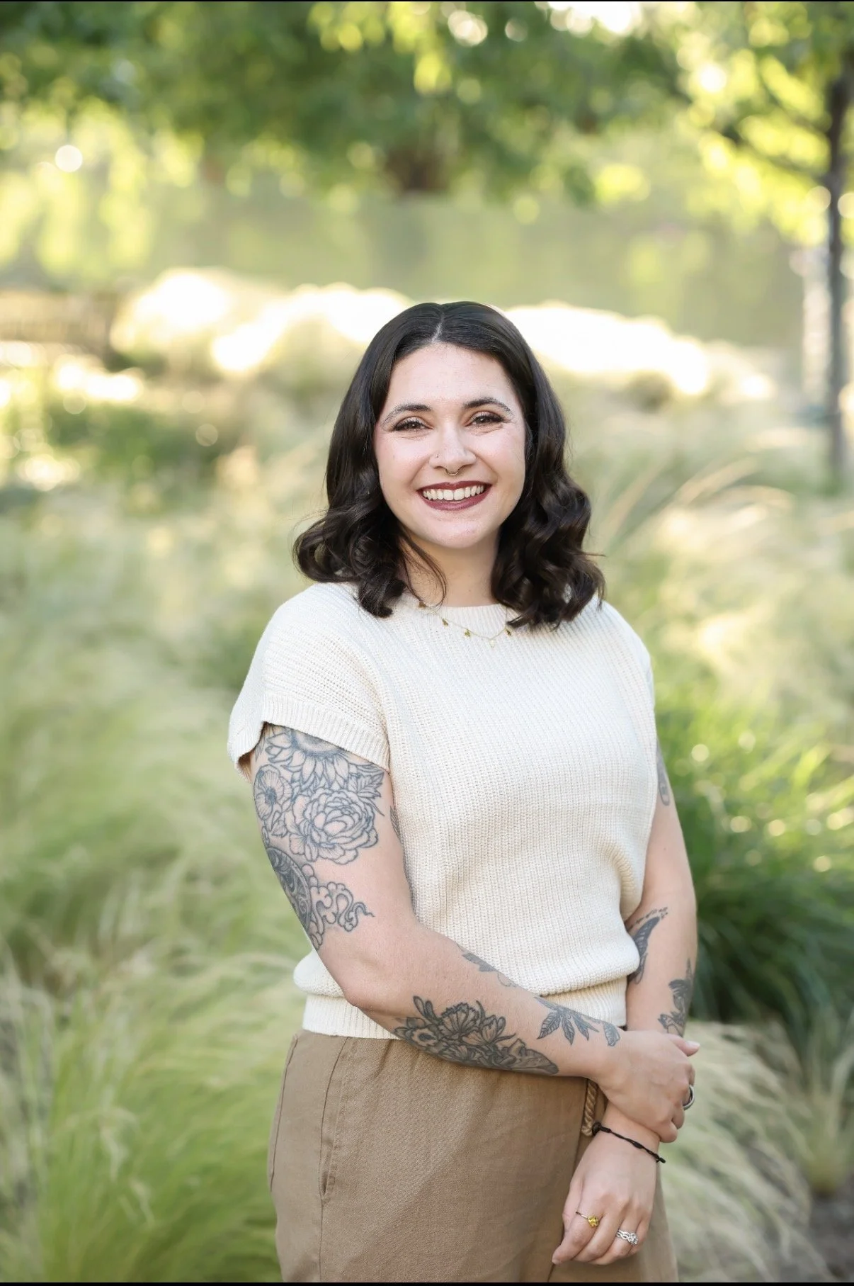 A woman with black, shoulder-length, curly hair smiling outdoors, wearing a cream-colored short-sleeve top and tan pants. She has tattoos on her arms and is standing in a green, sunlit park with trees and a pond in the background.