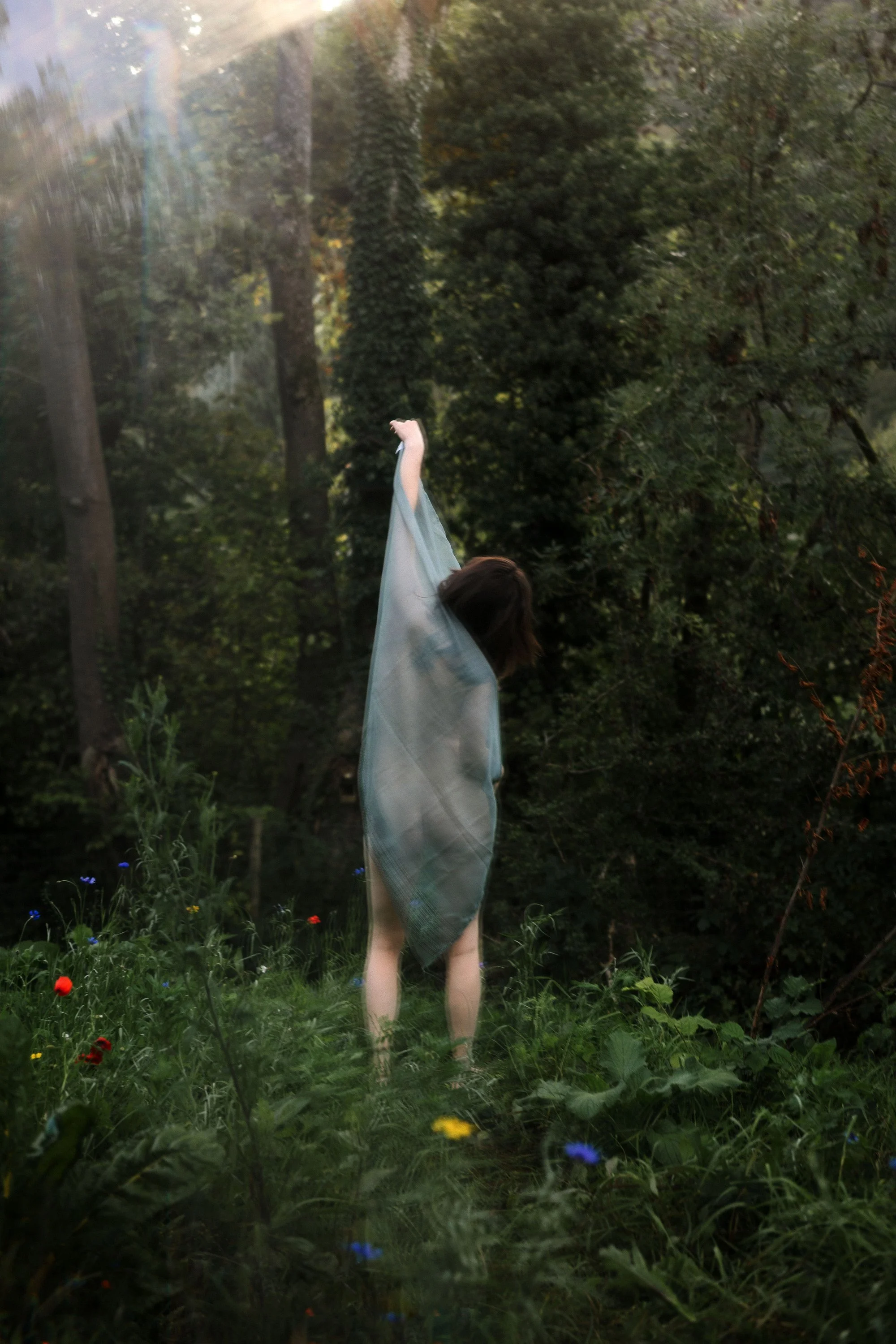 Une personne vue de dos dans une forêt, vêtue d'un tissu léger, levant un bras vers le haut, entourée de feuillages verts et de fleurs sauvages.