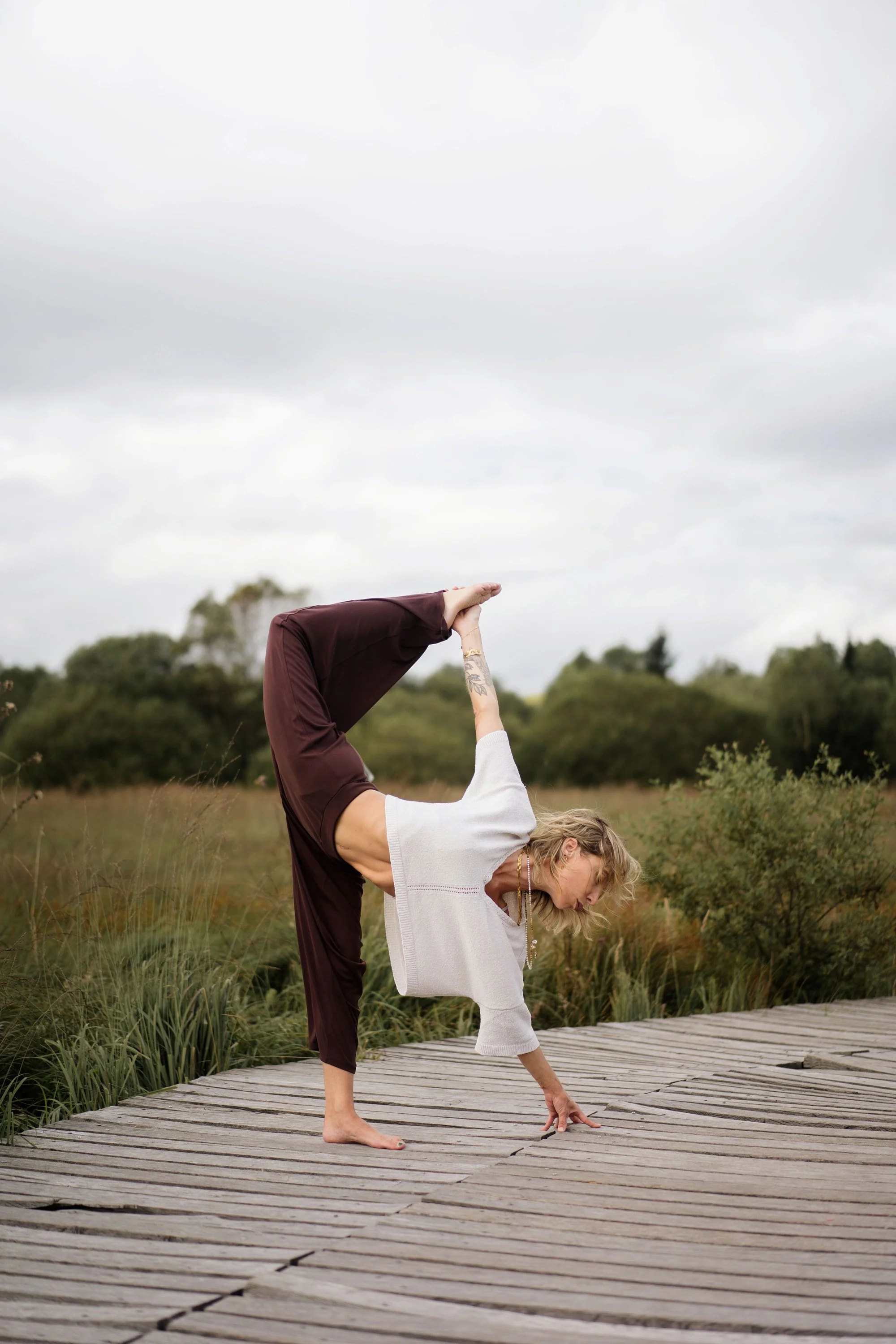 Une femme effectue une posture de yoga en équilibre sur une jambe, en tenant l'autre jambe derrière elle avec une main, dans un parc naturel. Elle porte un pull blanc et un pantalon marron, avec des cheveux bouclés et un tatouage visible sur le bras.