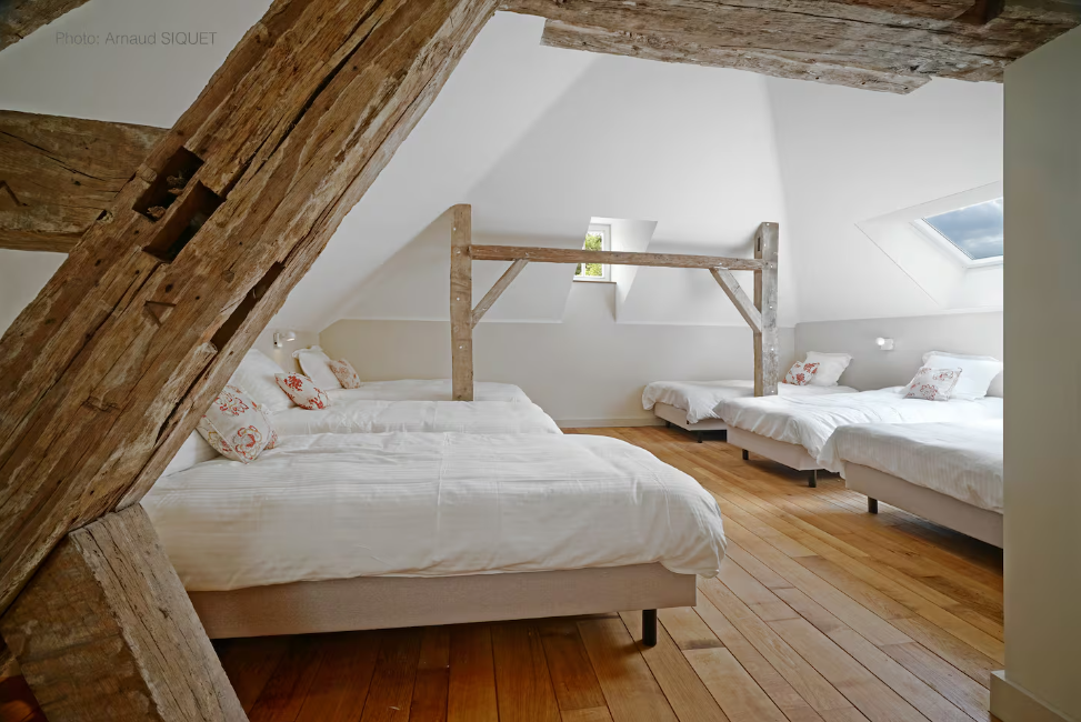 Chambre à coucher mansardée avec trois lits en blanc, une poutre en bois ancienne visible, fenêtre de toit et parquet en bois clair.