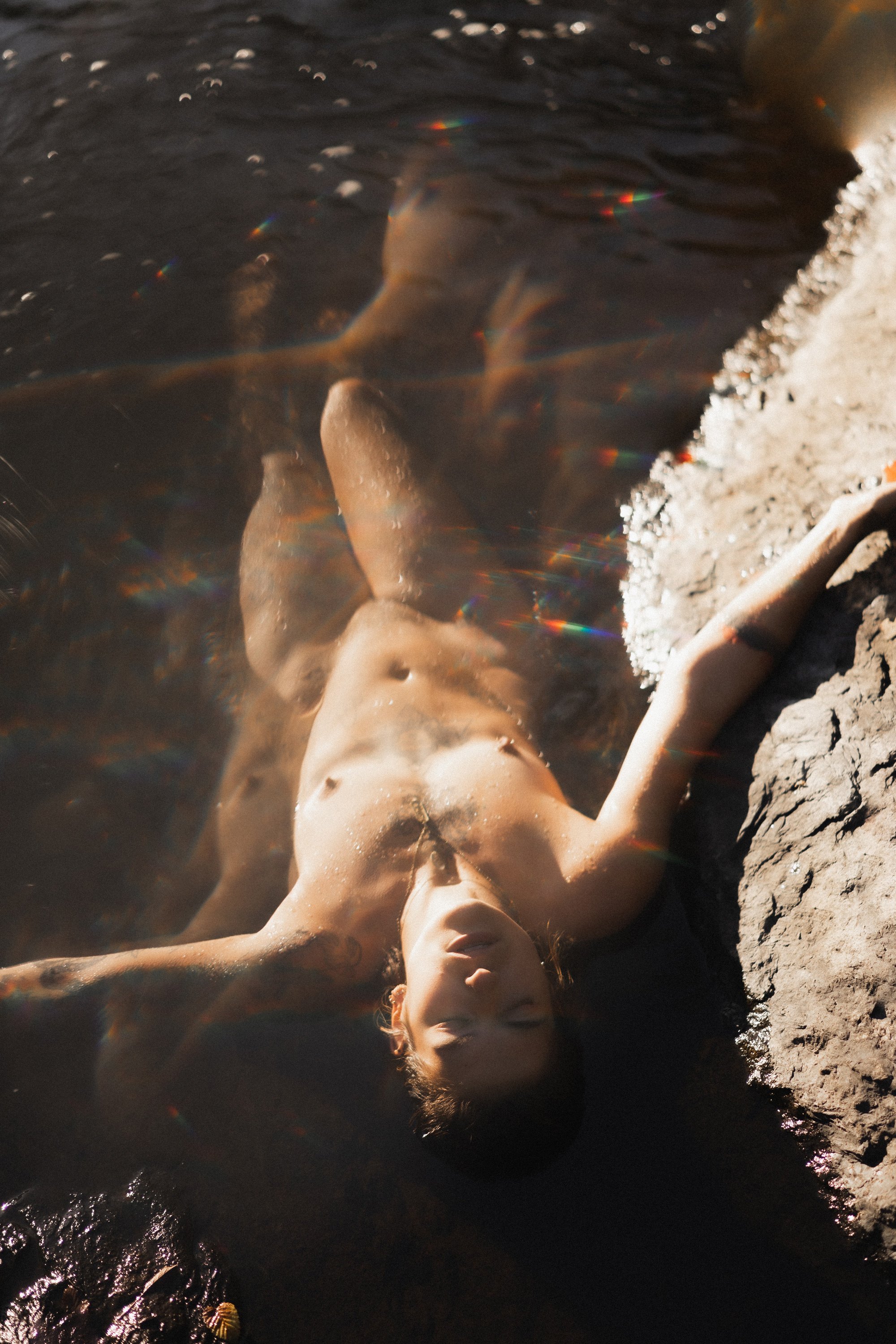 Une femme nue allongée sur le sable, partiellement immergée dans l'eau de mer, avec un ciel clair et le soleil brillant en arrière-plan.