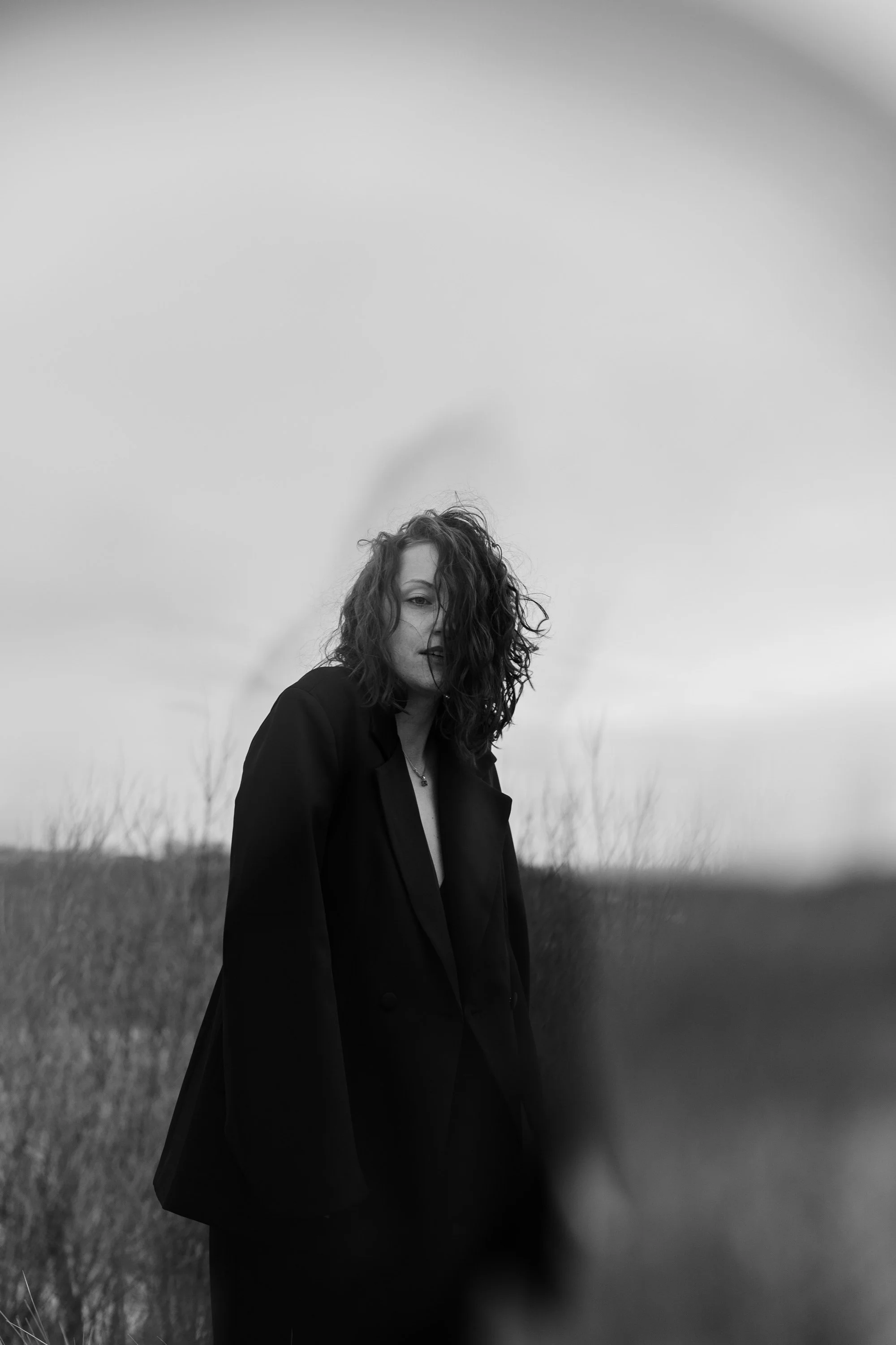 Une femme aux cheveux bouclés porte un manteau noir dans un champ, avec un ciel couvert en arrière-plan, en noir et blanc.