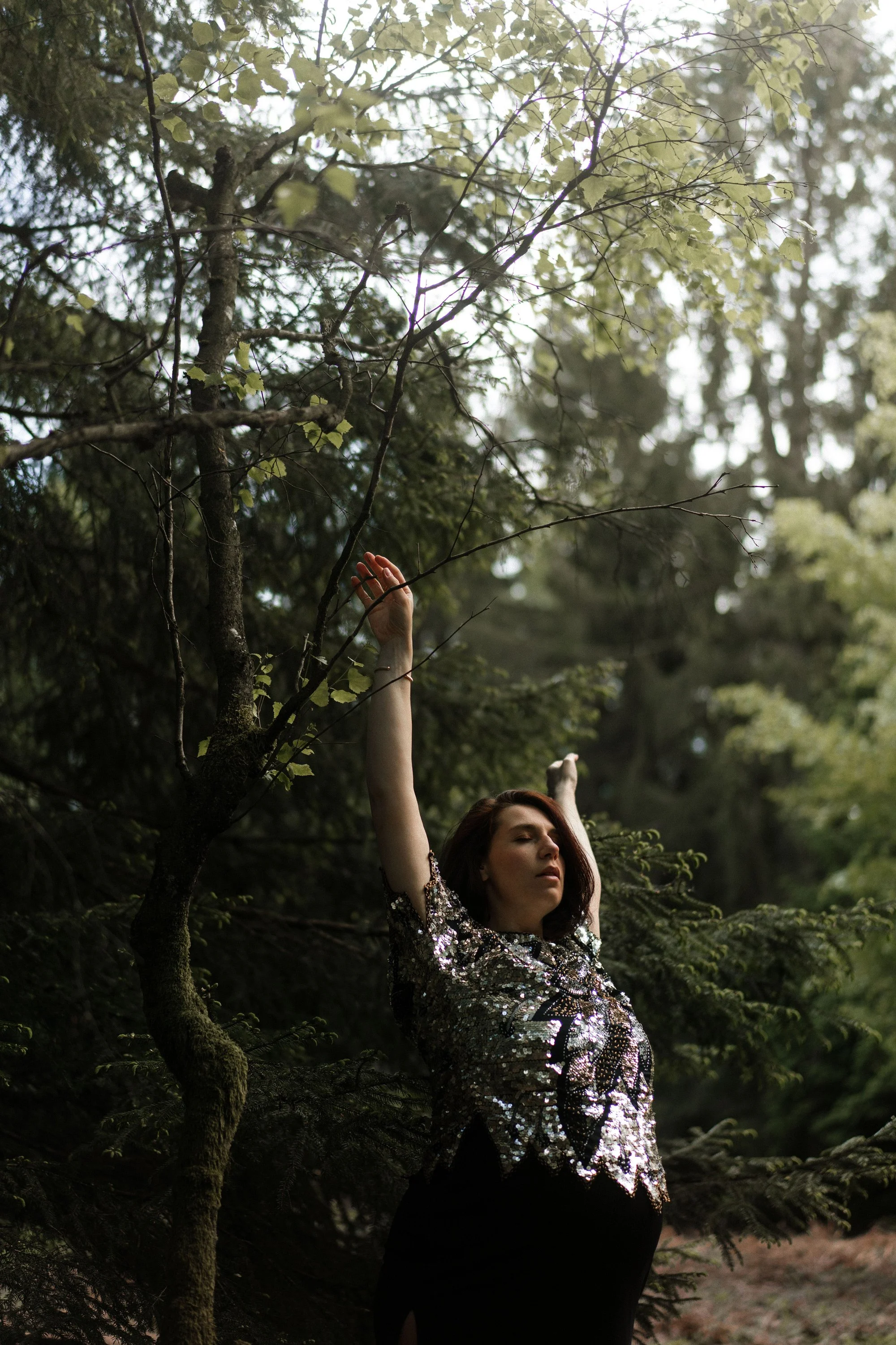 Une femme avec une robe à sequins noires et argentées se tient dans une forêt, les bras levés, avec des arbres verts en arrière-plan.