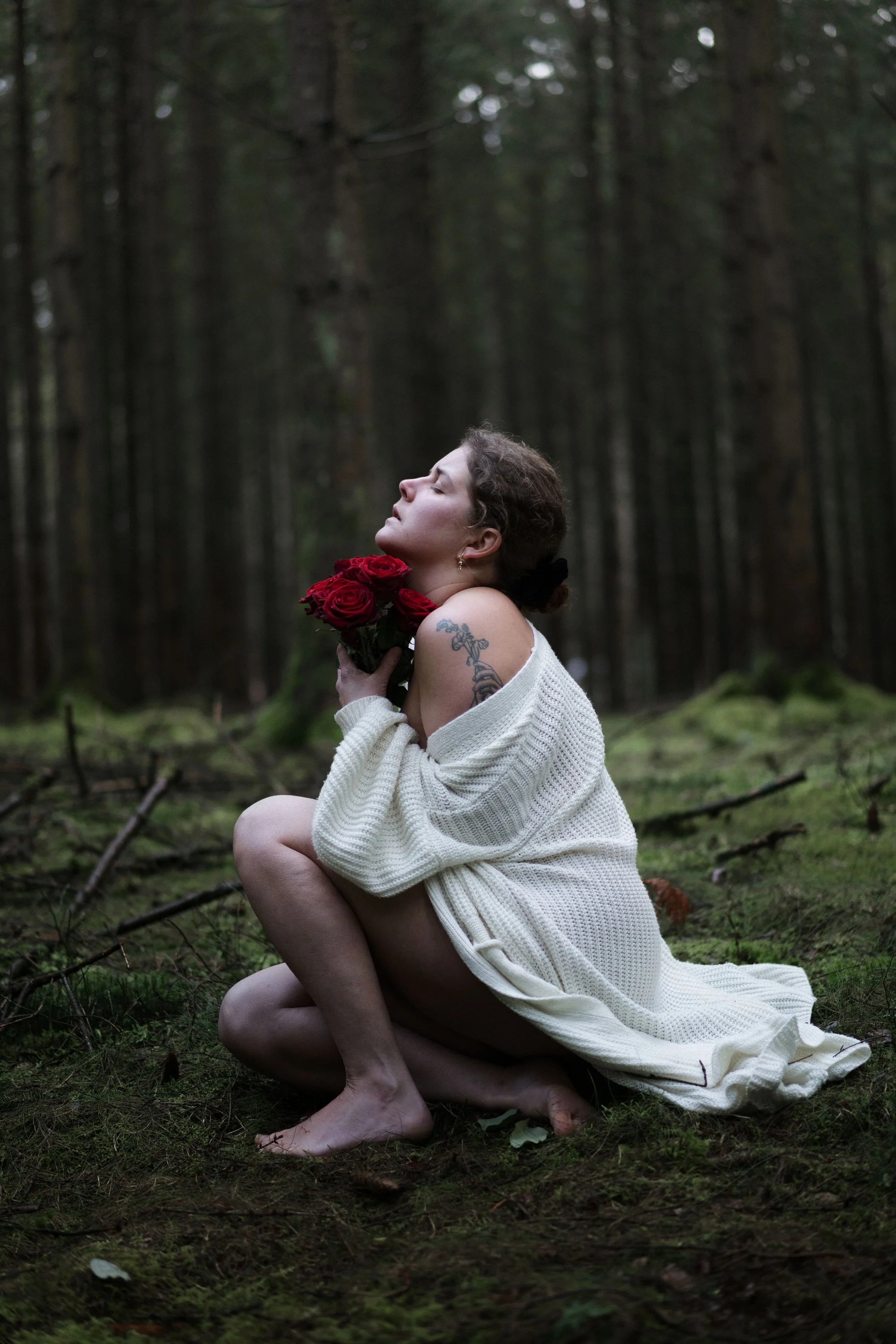 Une femme nue, assise dans une forêt, tenant un bouquet de roses rouges près de son visage, avec une expression de sérénité.