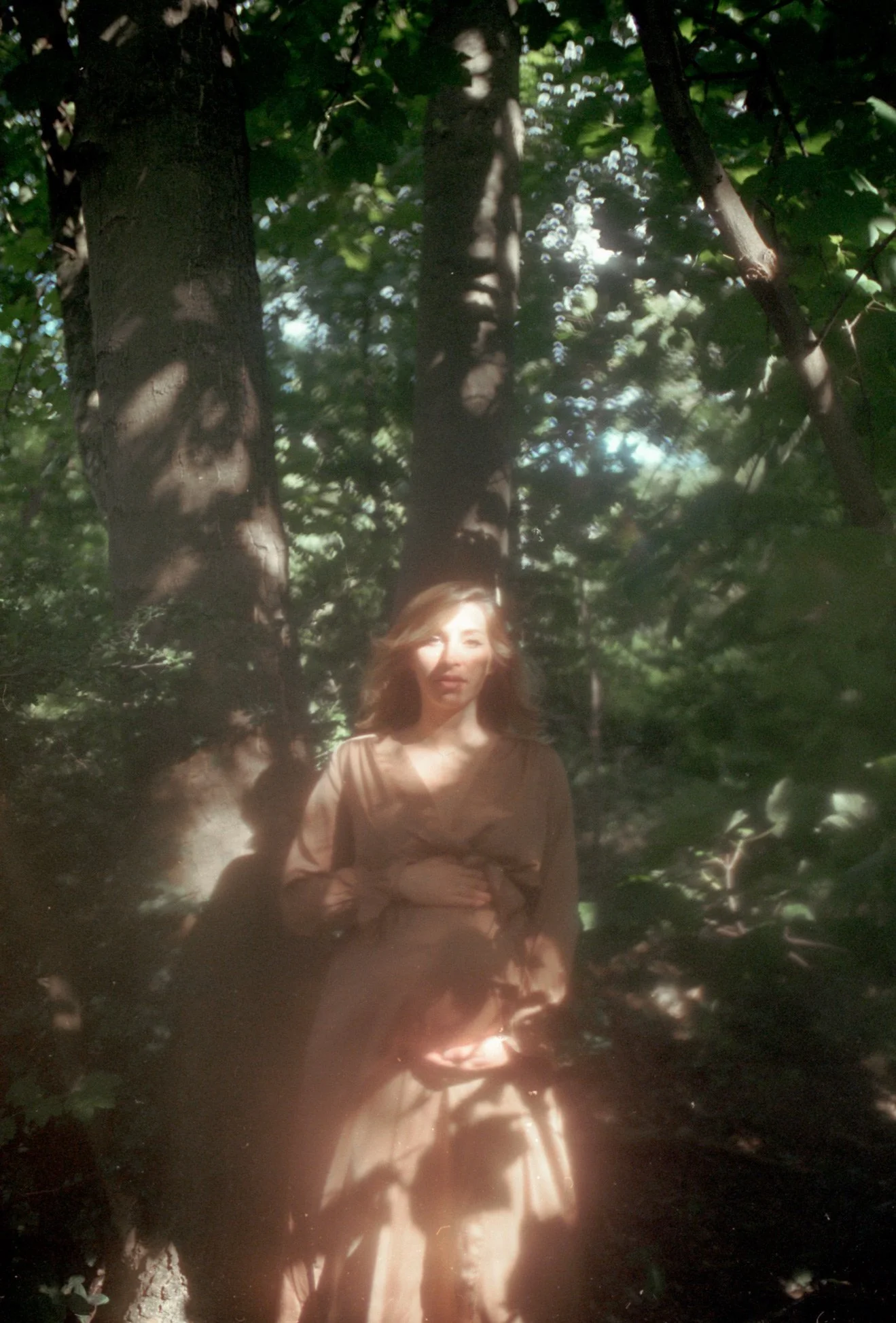 Une femme avec des cheveux roux, portant une robe beige, se tient dans la forêt parmi les arbres, avec des ombres de feuilles projetées sur elle et autour d'elle.