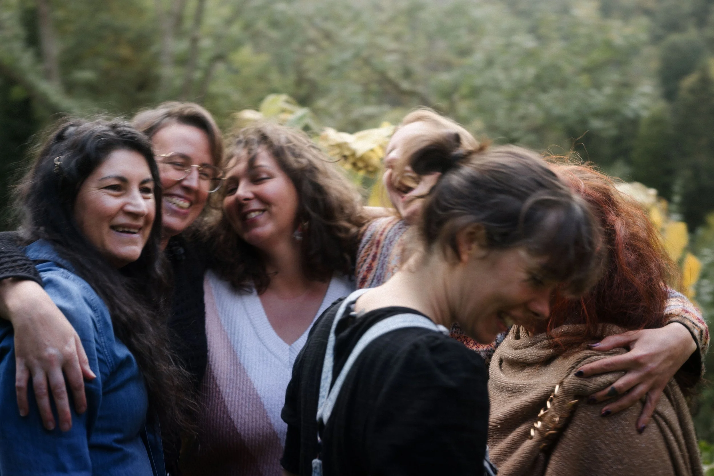 Un groupe de femmes souriantes se tenant par les épaules en cercle dans un environnement extérieur verdoyant.