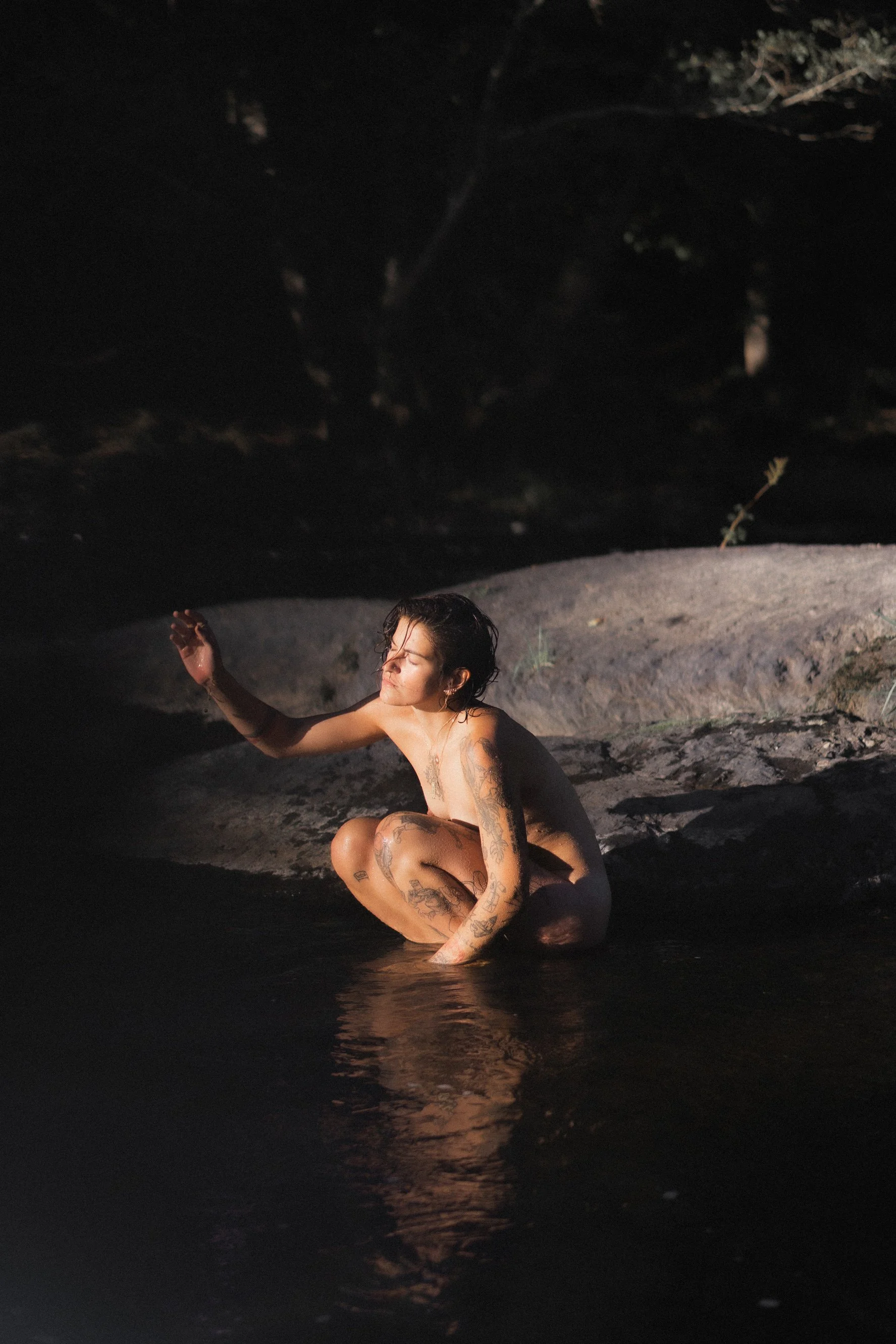 Une femme nue assise dans une rivière ou un lac la nuit, entourée de rochers, avec des tatouages sur ses bras, ses jambes et son torse, les yeux fermés, sous la lumière douce de l'eau.