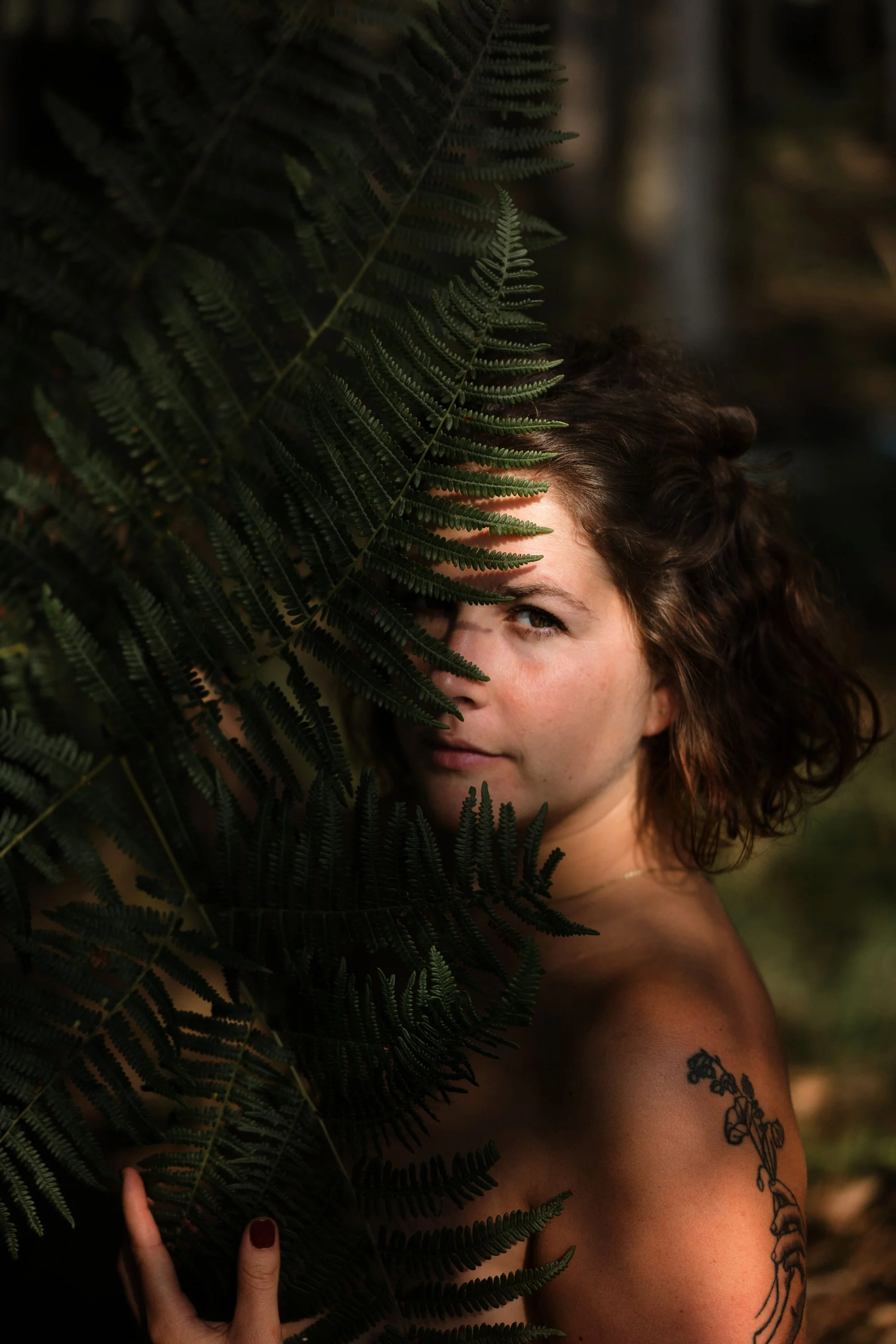 Femme avec des tatouages, tenant des fougères, regardant la caméra dans un environnement naturel.