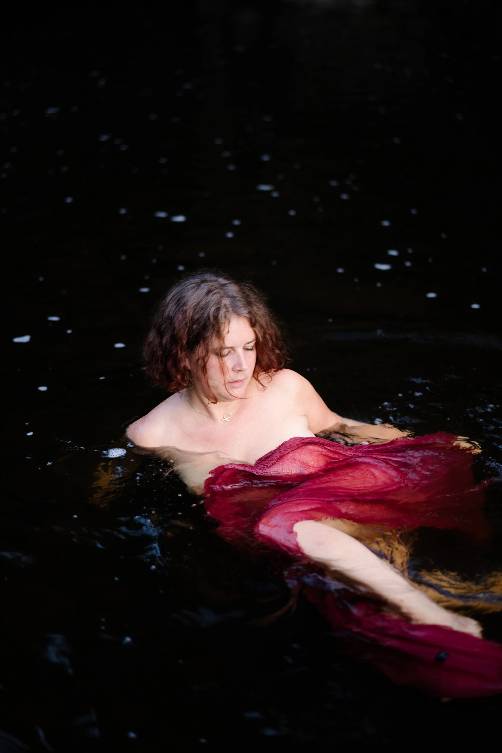 Une femme nue émerge de l'eau sombre, portant une robe rouge. Elle a des cheveux bouclés et regarde vers le bas.