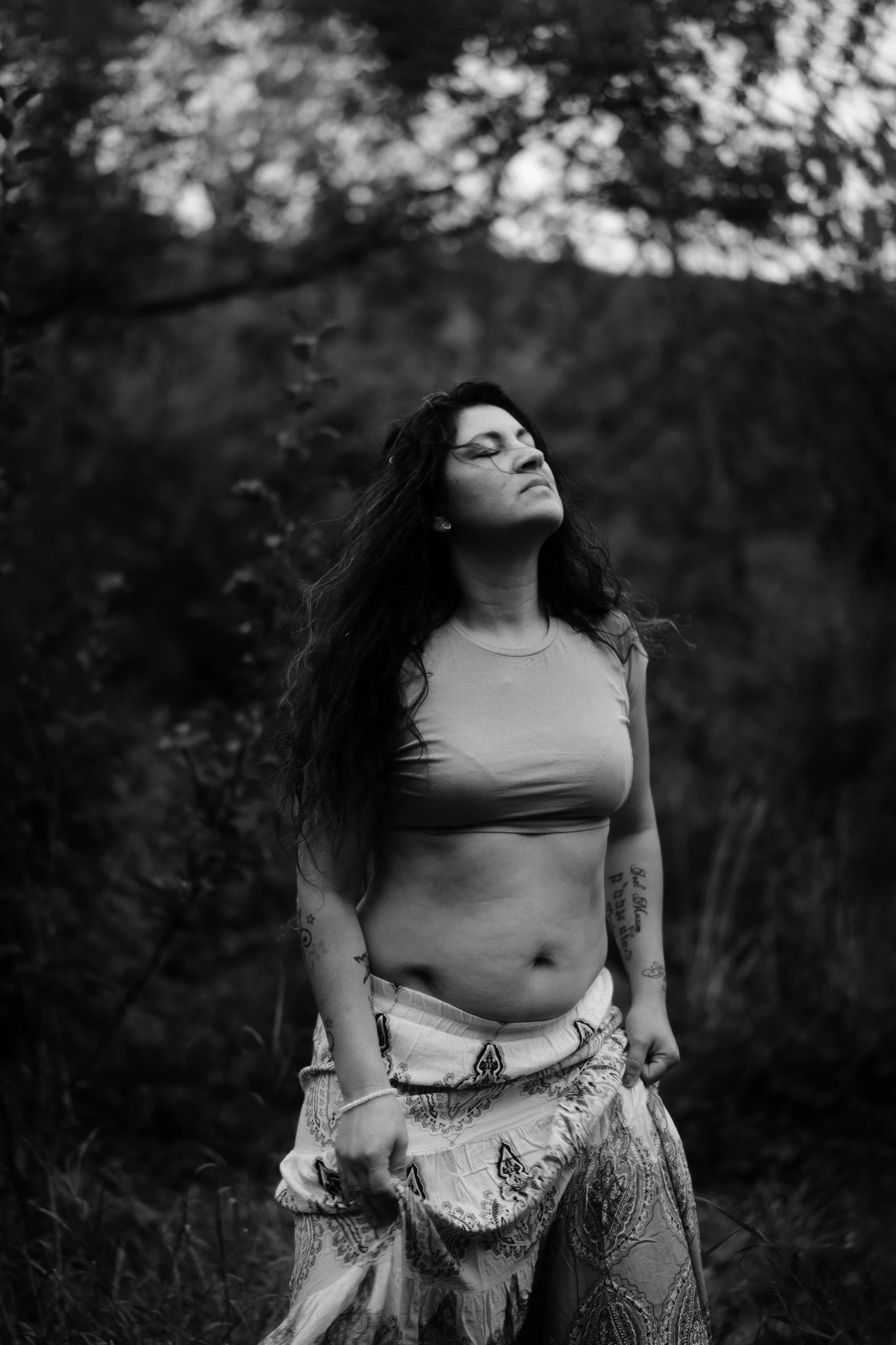 Une femme noire avec des tatouages, portant un crop top et une jupe longue et floquée, se tient dans un environnement naturel en plein air, en noir et blanc.