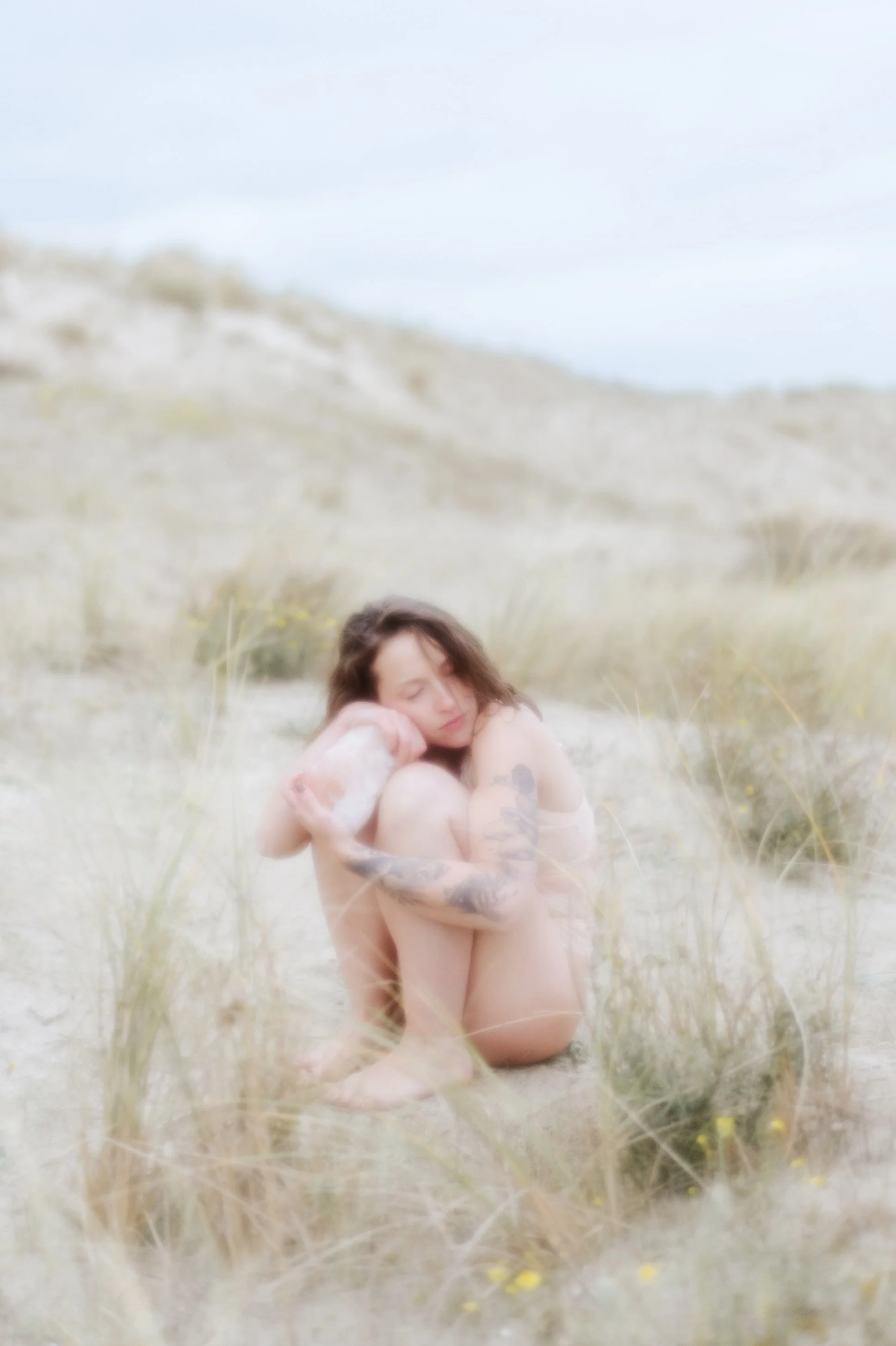 Une femme nue avec des tatouages, assise à genoux dans un champ d'herbes, la tête inclinée et fermée, dans un paysage naturel avec des dunes et un ciel clair.