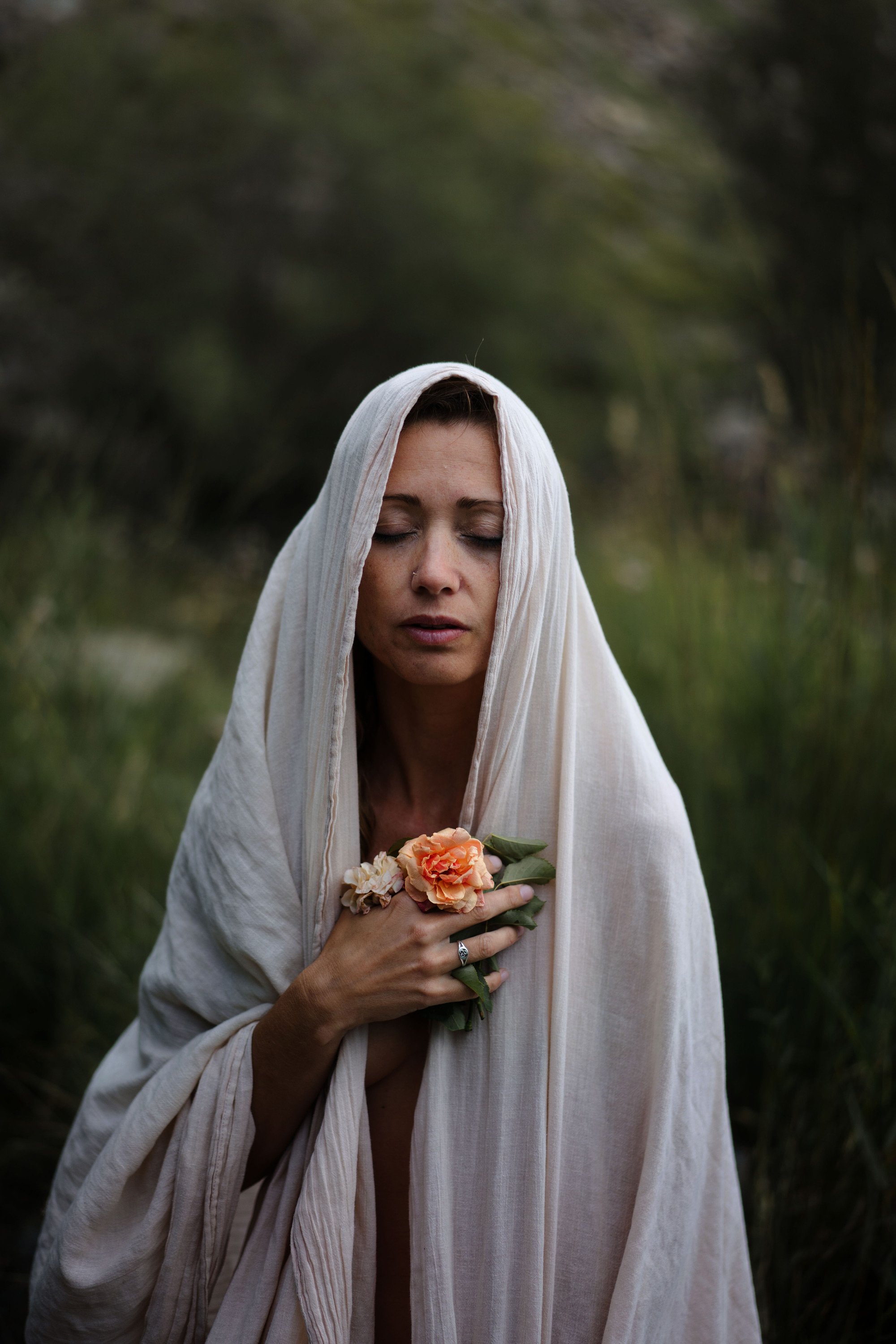 Une femme avec les yeux fermés, portant un voile blanc sur la tête, tenant un bouquet de fleurs, dans un environnement naturel avec des arbres flous en arrière-plan.