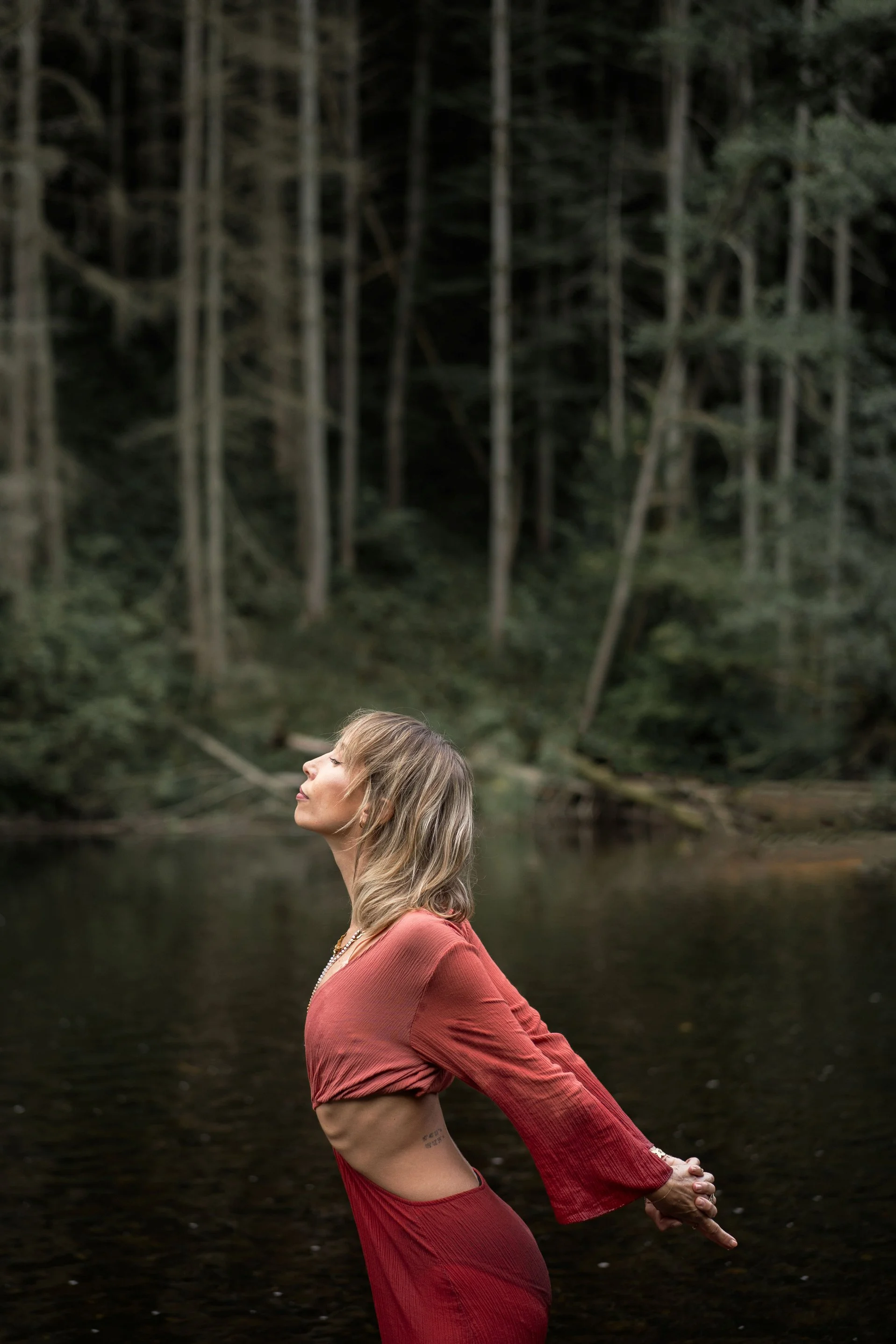 Une femme en posture de yoga dans une nature forêt, les yeux fermés, le visage détendu, portant une robe rouge oranger, à côté d'une rivière ou d'un lac, avec des arbres en arrière-plan.