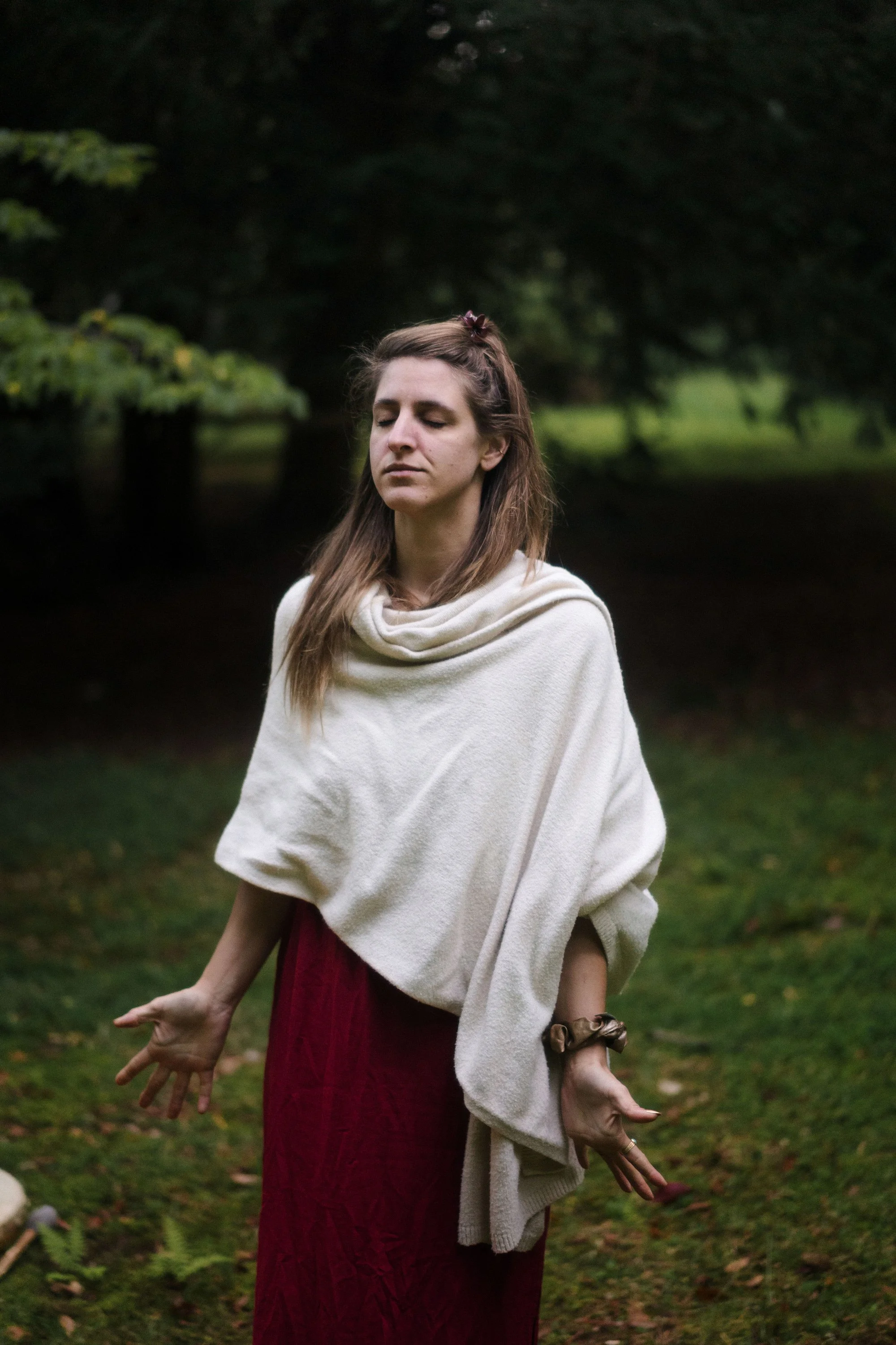 Une femme avec les yeux fermés, portant une robe rouge et une écharpe beige, se tenant dans un cadre naturel verdoyant avec de l'eau en arrière-plan.