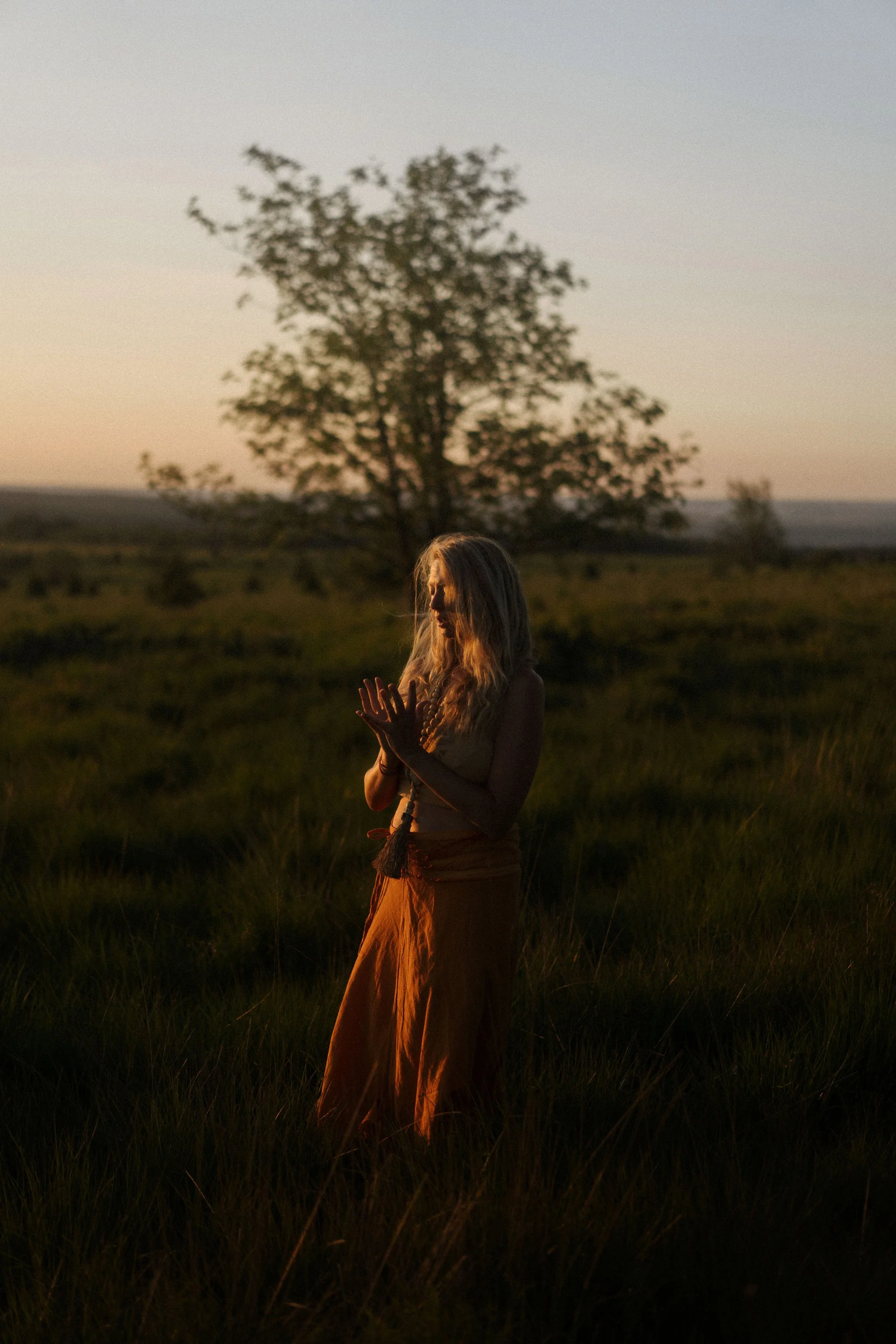 Une jeune femme avec des cheveux longs et bouclés, portant une jupe longue de couleur orange, se tenant dans un champ avec de l'herbe haute, au coucher du soleil ou au lever du soleil, avec un arbre en arrière-plan.