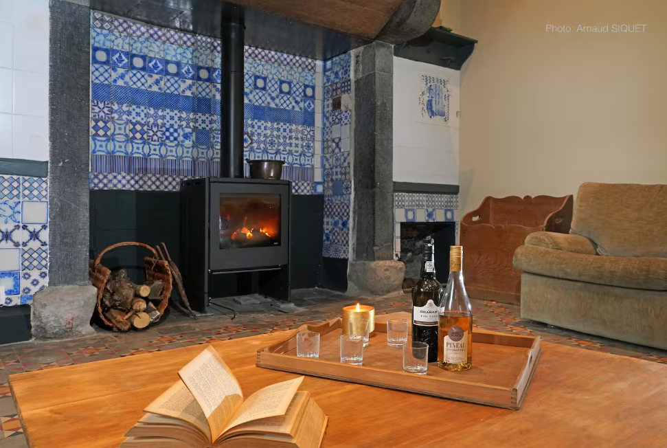 Salon avec cheminée en pierre, fauteuil, canapé, table en bois avec livres, bouteilles d'alcool, bougie allumée, et tapis de sol.