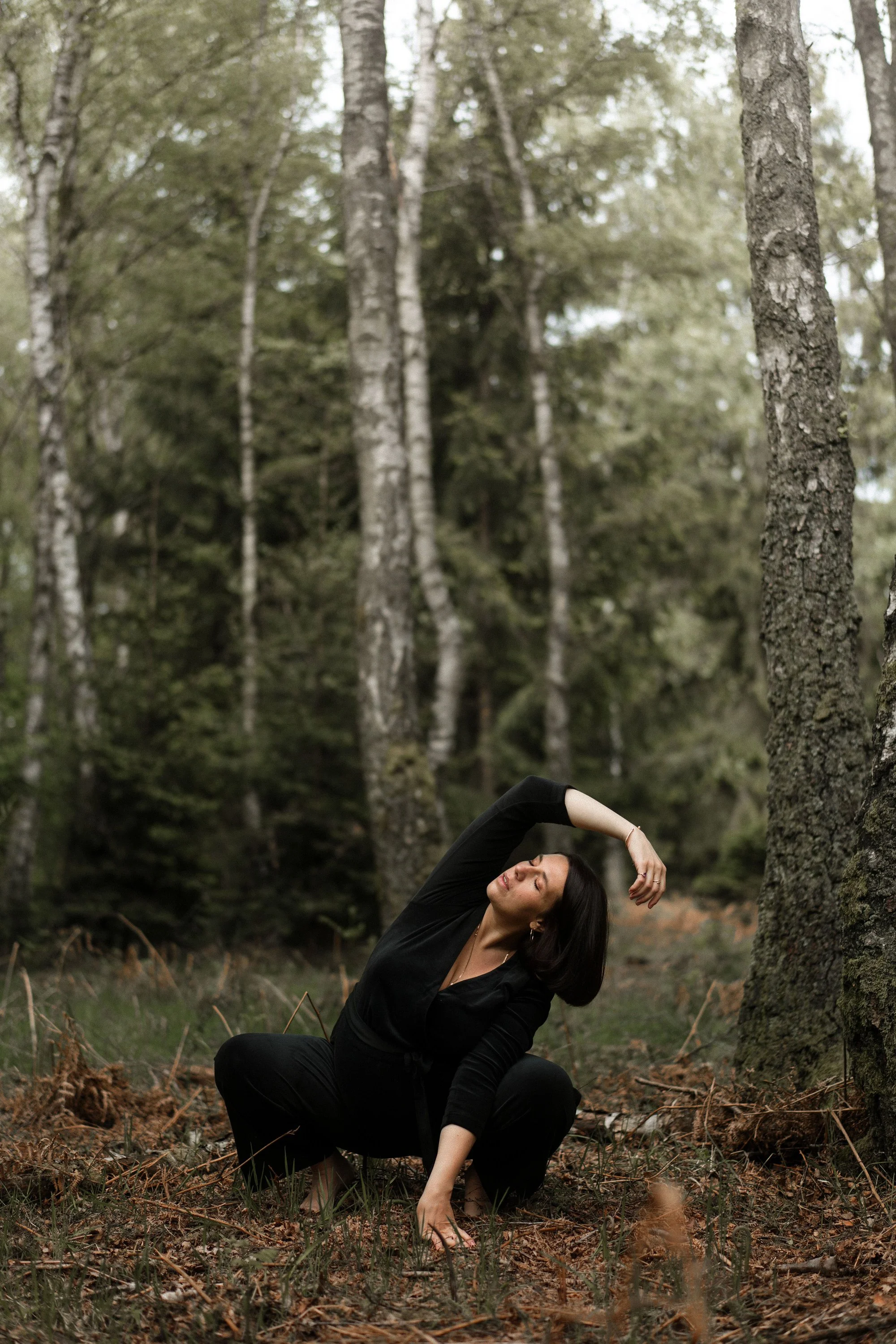 Une femme en position de yoga dans une forêt dense, entourée d'arbres et de feuilles au sol.