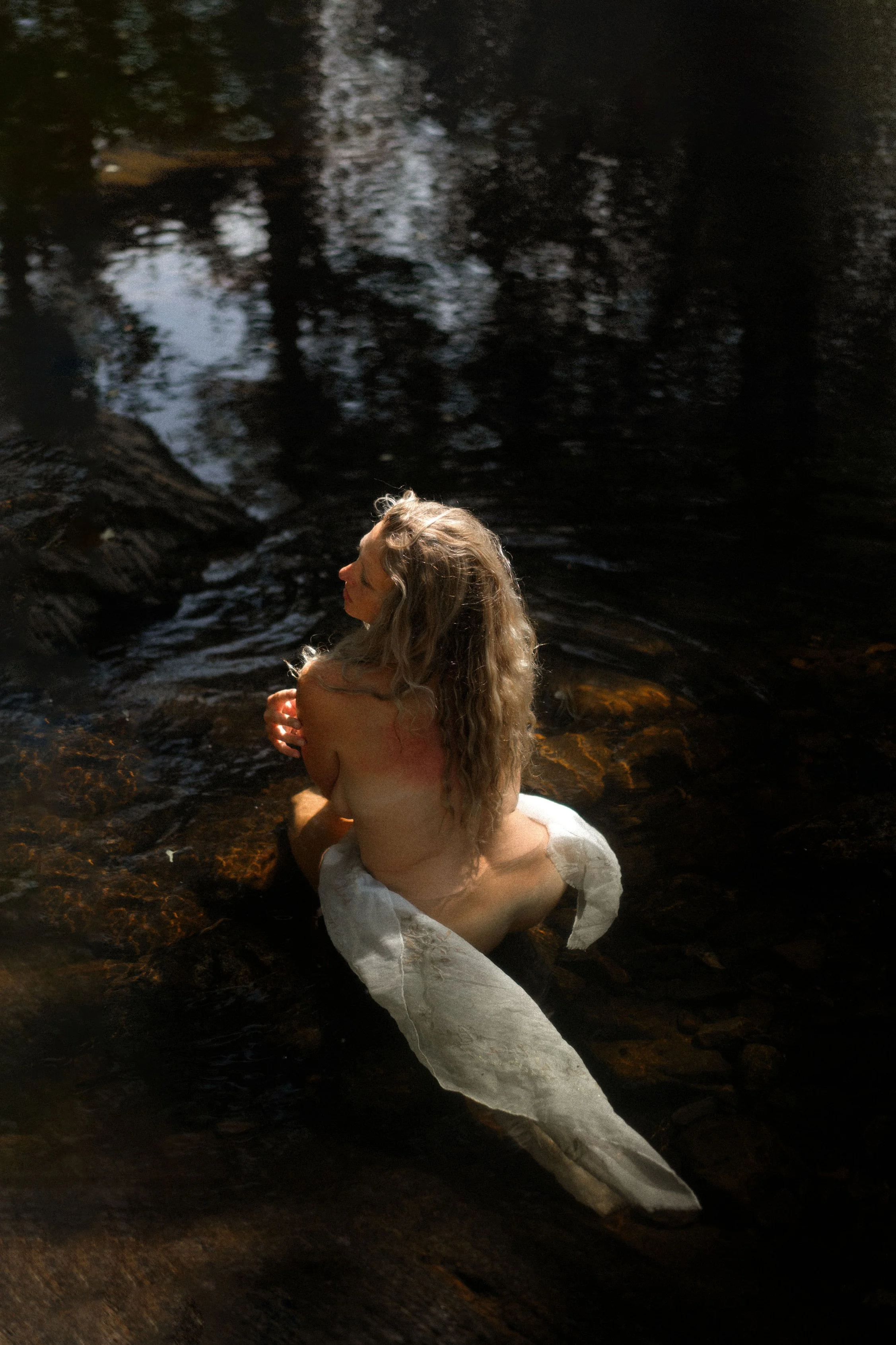 Une femme nue assise dans une rivière, portant une longue jupe blanche et ses longs cheveux blonds cascades sur ses épaules, avec une surface d'eau sombre et réfléchissante en arrière-plan.