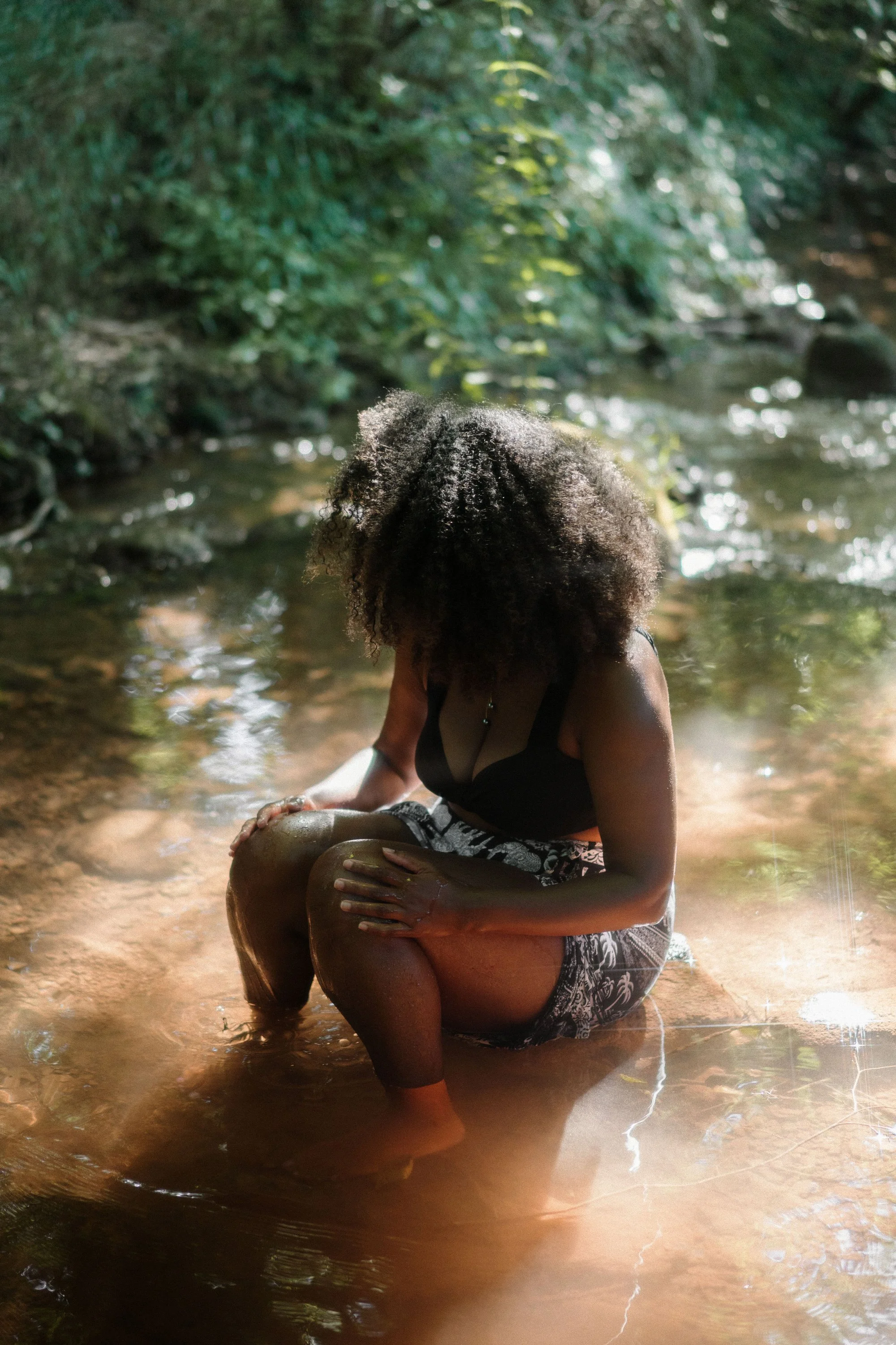 Une femme assise dans une rivière naturelle entourée de végétation verte dense, portant un maillot noir et des shorts, avec des cheveux bouclés.