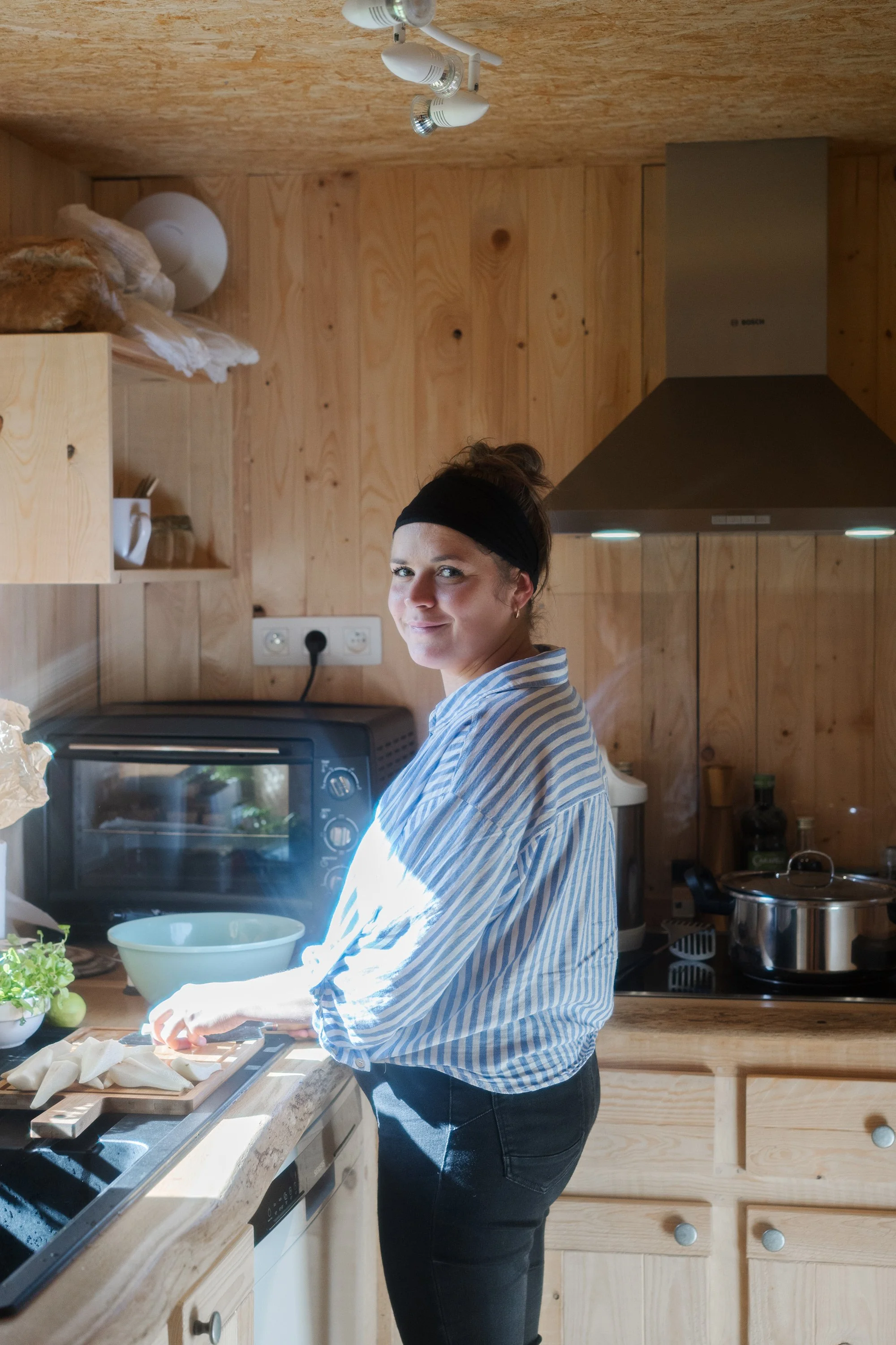 Une femme en chemise à rayures, coiffée d'un bandeau noir, prépare des aliments dans une cuisine en bois, devant un four. Elle sourit en regardant l'appareil photo.