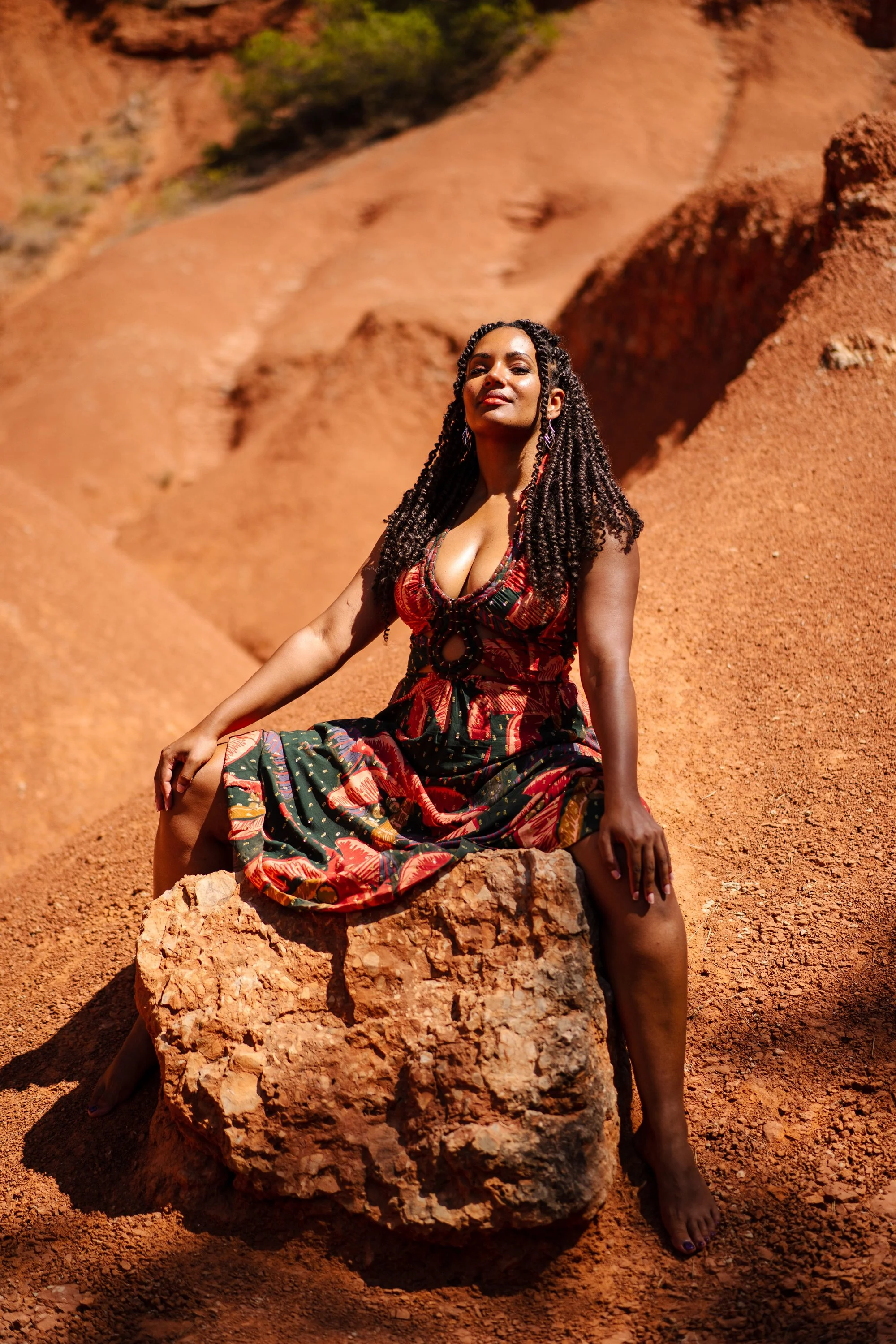 Une femme assise sur un rocher dans un paysage rocheux rouge, portant une robe colorée avec des motifs floraux, sous un ciel ensoleillé.