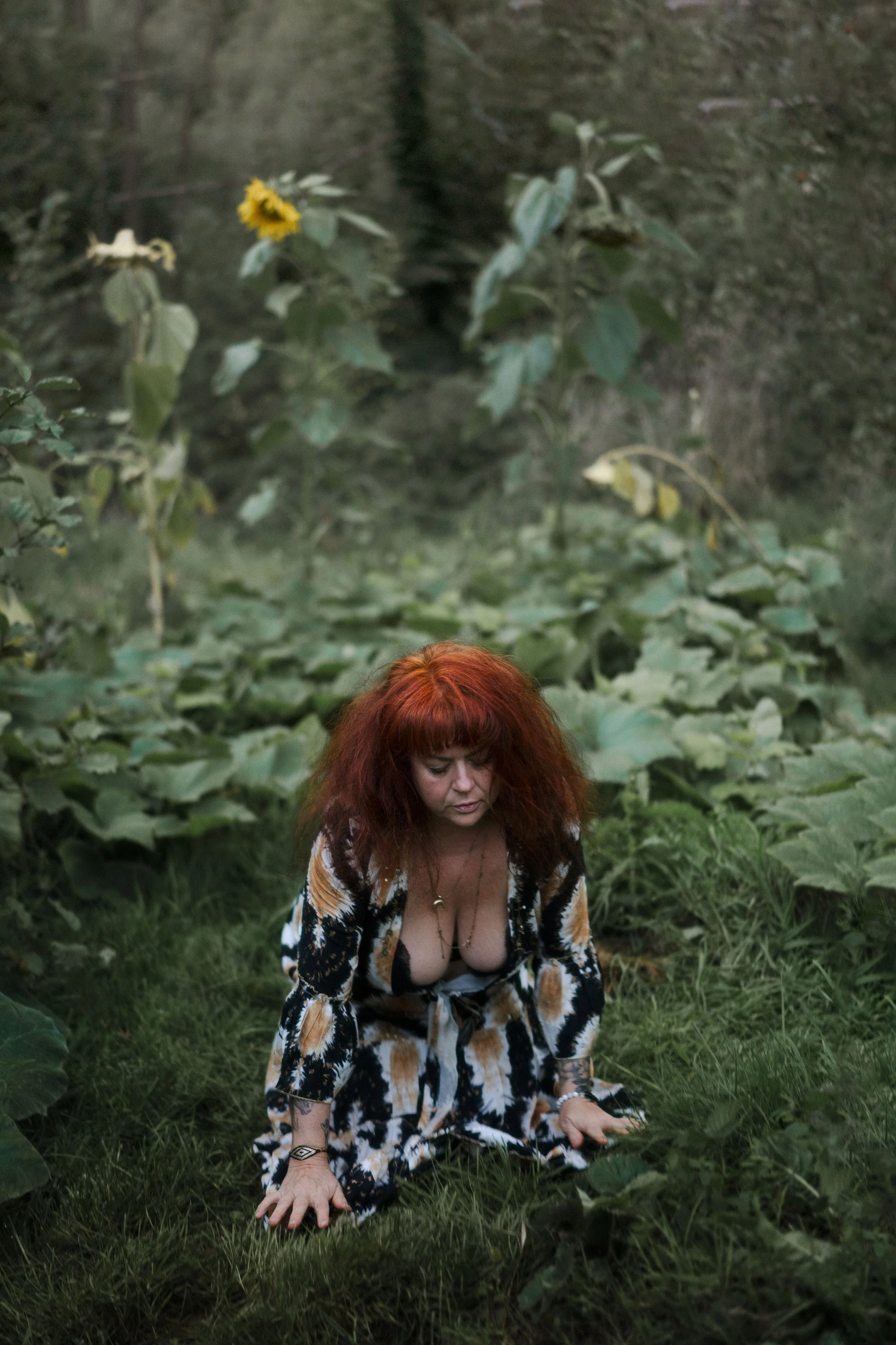 Une femme aux cheveux roux bouclés, vêtue d'une robe ou d'un manteau à motif animal, est à genoux dans une végétation verte dense. Son regard est dirigé vers le sol, dans un environnement naturel, près d'une branche ou d'une feuille.
