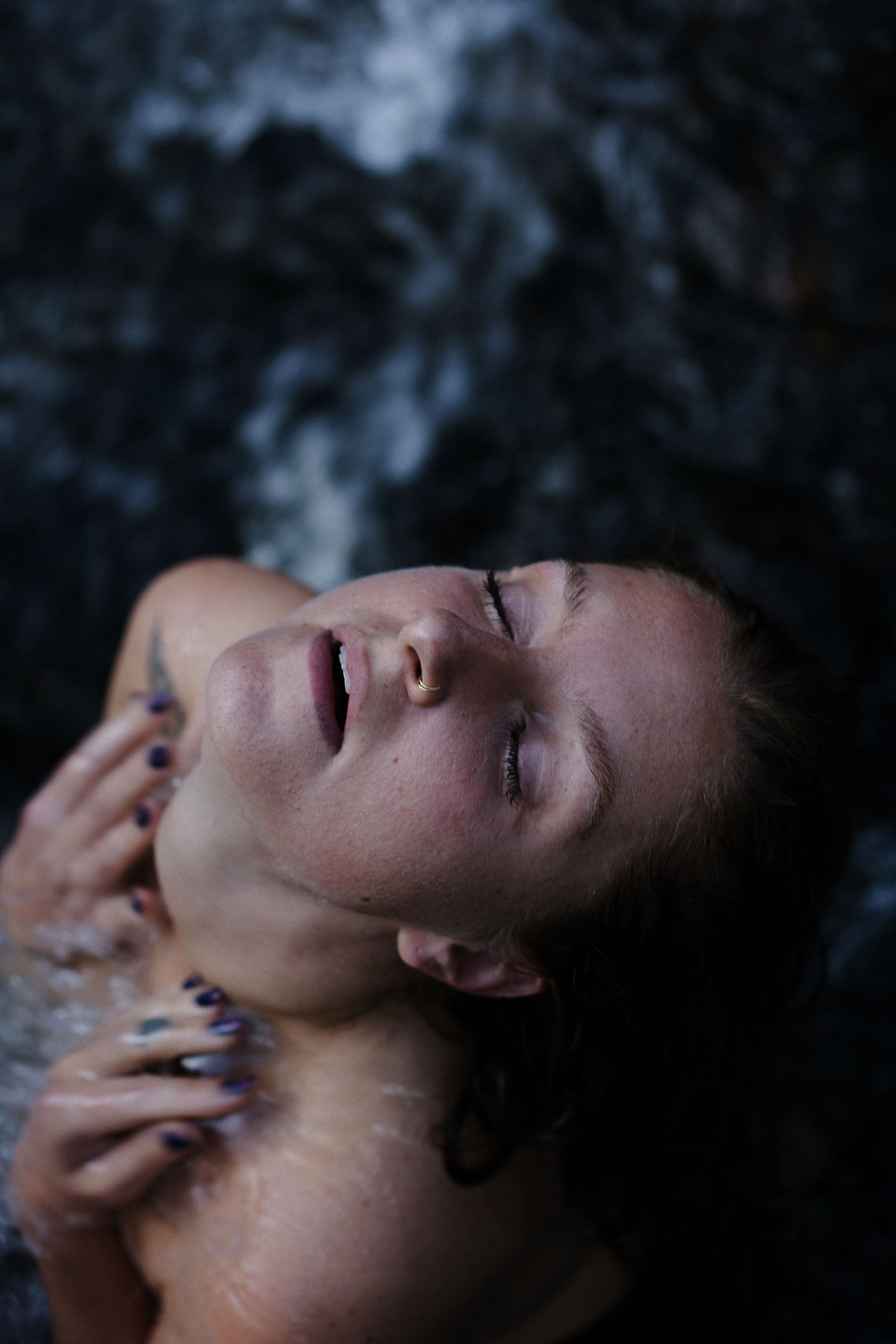 Jeune femme avec des yeux fermés, portant un anneau au nez, nageant dans l'eau, cheveux humides, peau éclatante.