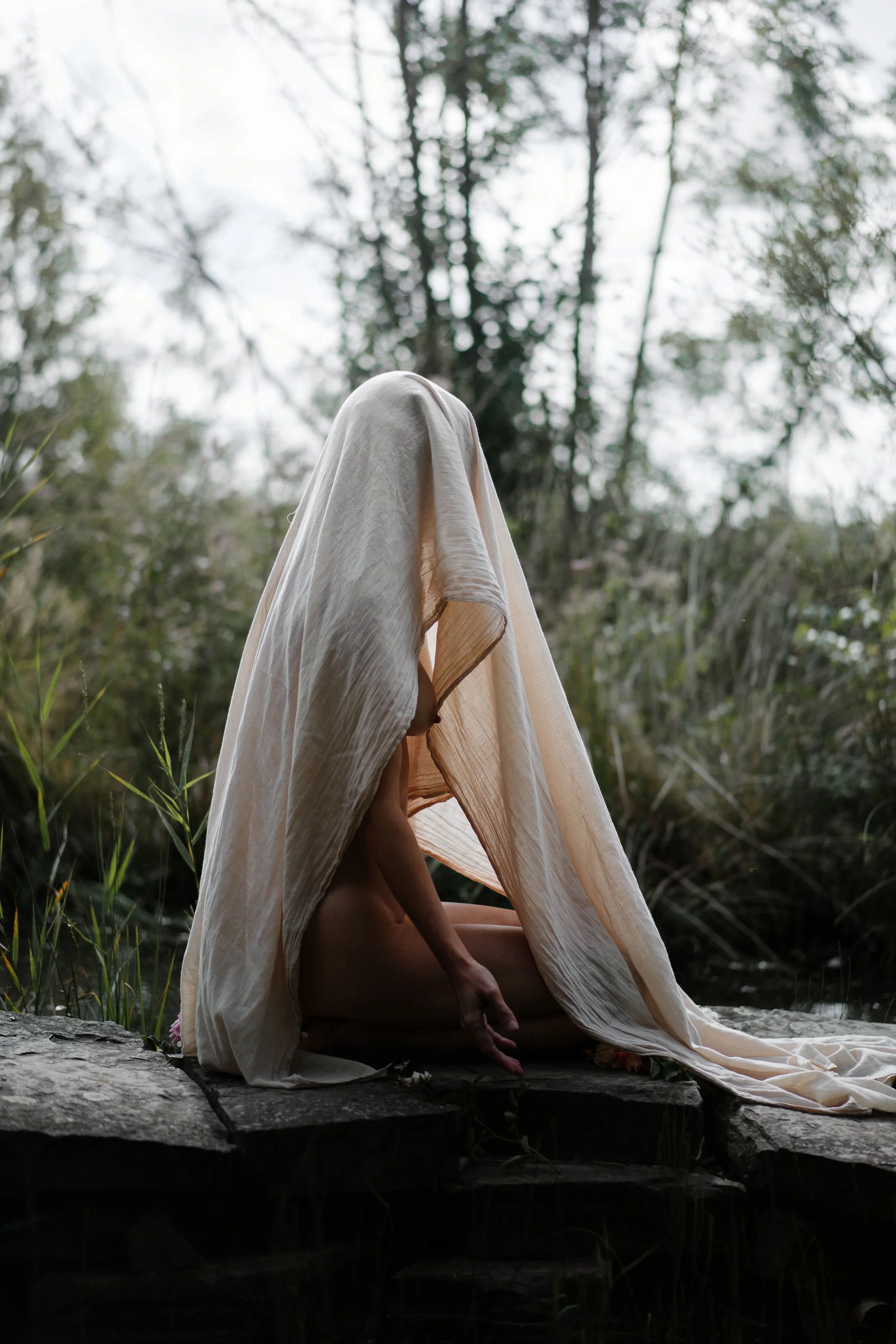 Une personne est assise en méditation dans la nature, cachée derrière un tissu léger beige qui forme une tente ou un abri, dans un environnement forestier.