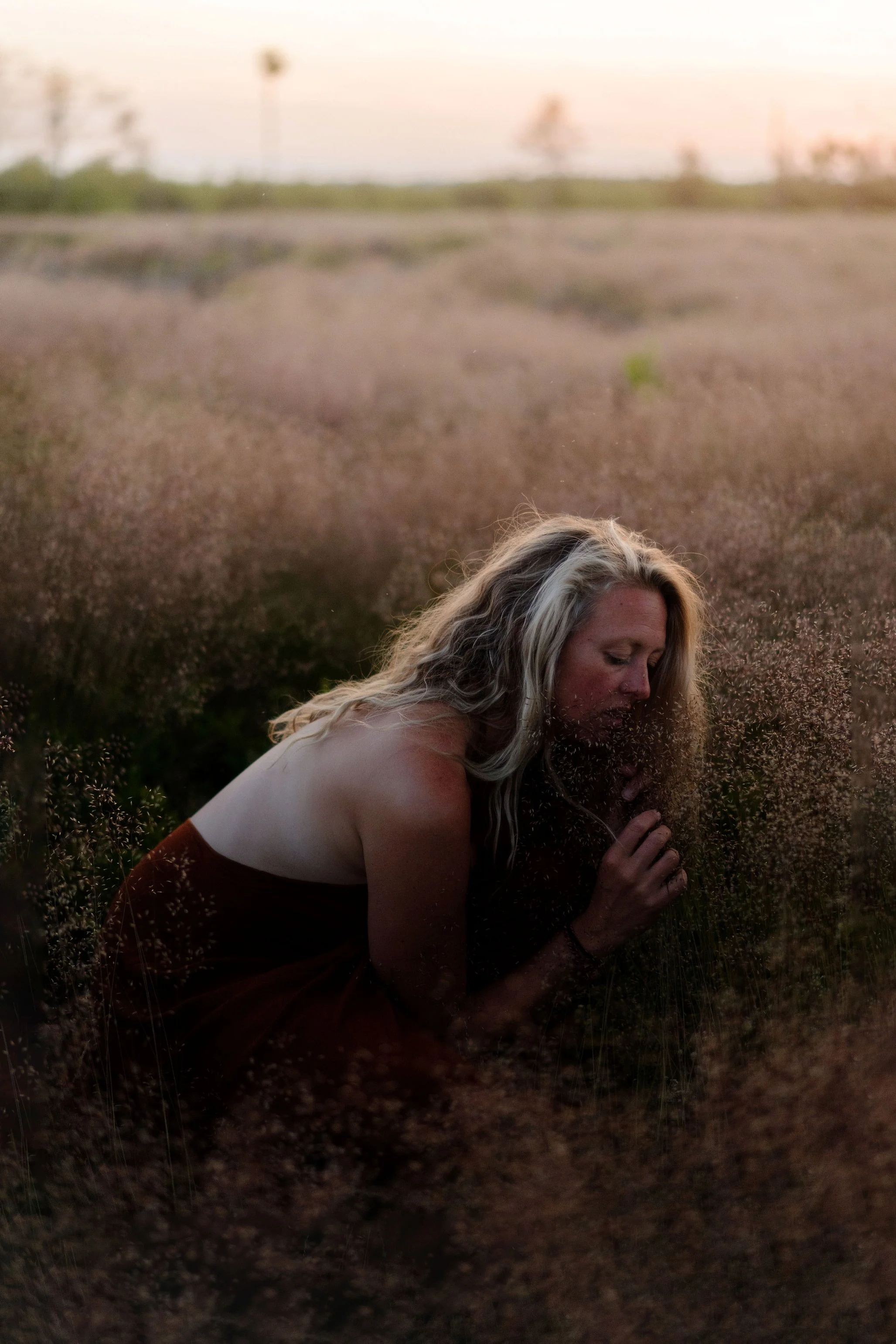 Femme aux cheveux longs et blonds, enroulée dans des vêtements sombres, dans un champ de plantes petites et fines, sous la lumière du coucher de soleil.
