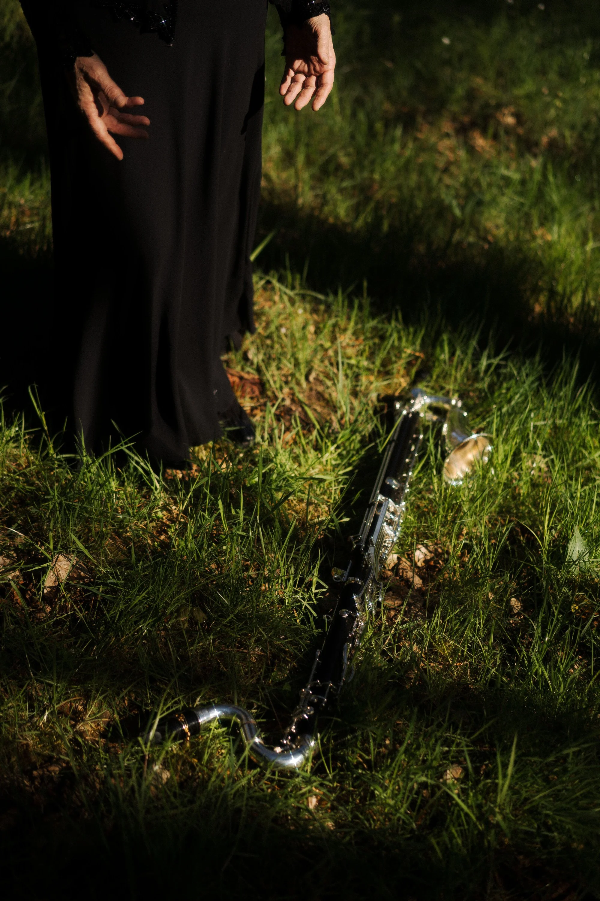Une personne habillée en noir, debout sur de l'herbe, avec un clarinette renversée sur le sol devant elle.