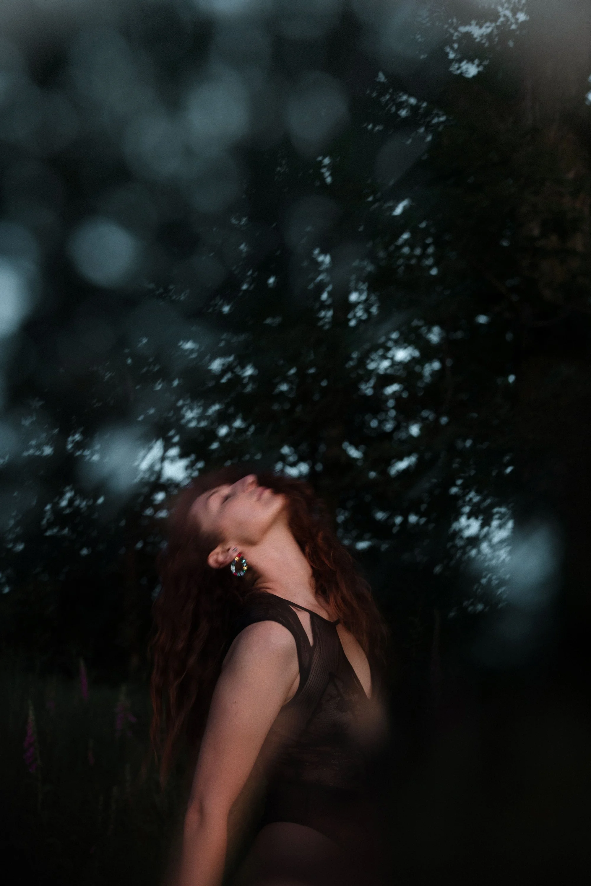 Une femme avec de longs cheveux roux, portant des boucles d'oreilles colorées, pose en extérieur avec des arbres en arrière-plan lors du crépuscule ou de la nuit.