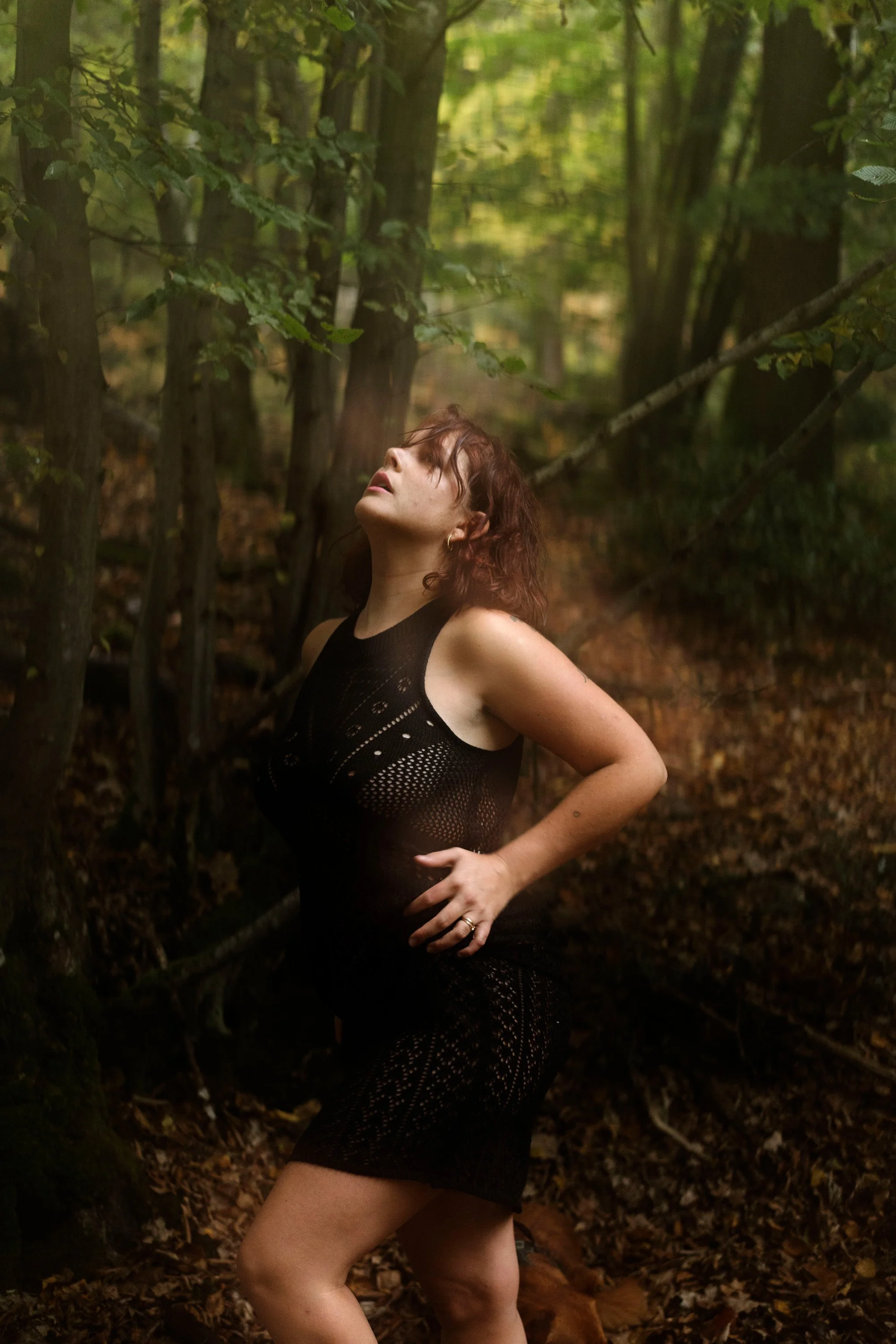Une femme avec des cheveux bruns, portant une robe noire en dentelle, pose dans une forêt avec des arbres verts et un sol couvert de feuilles.