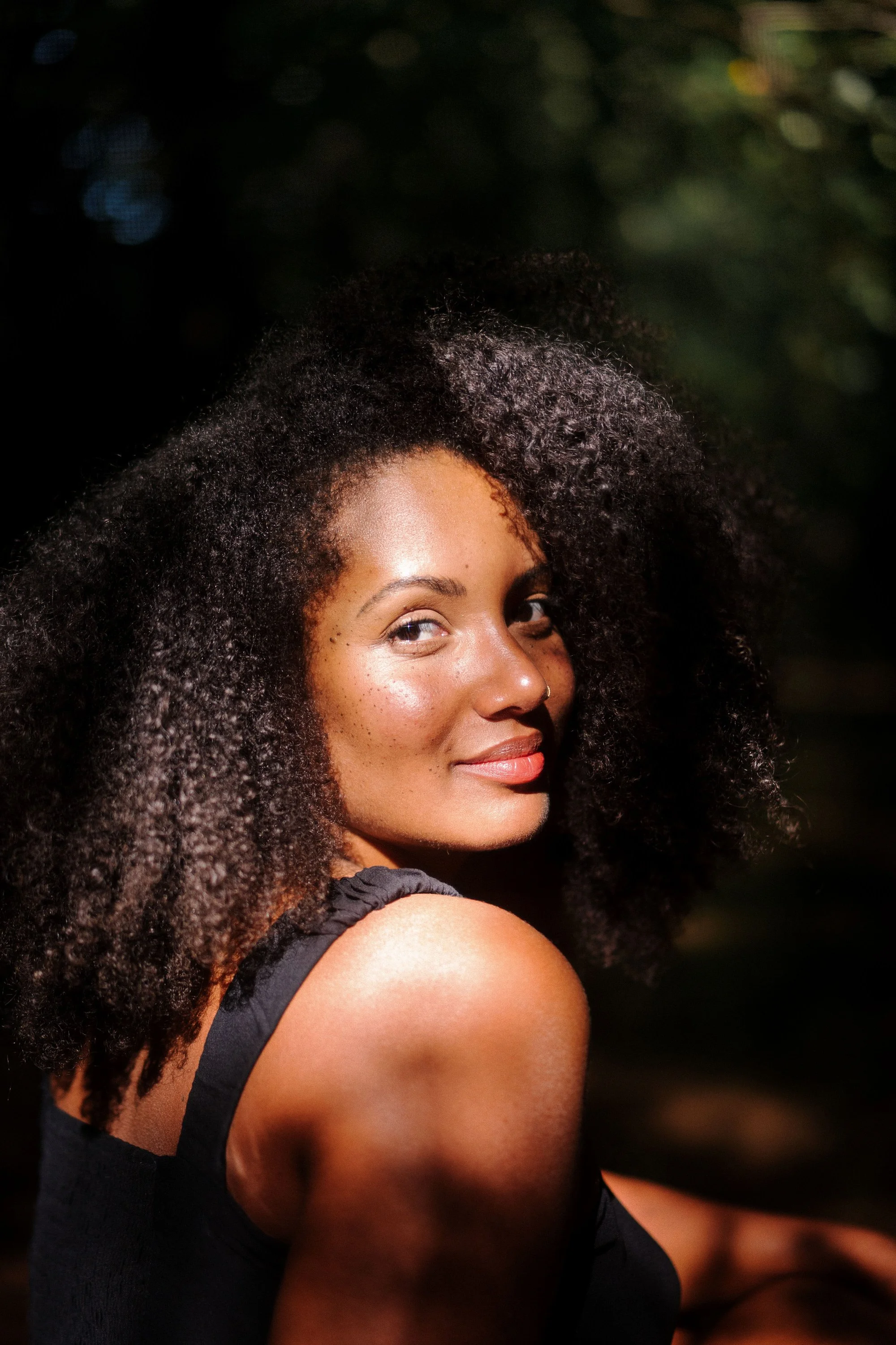 Une femme souriante avec de longs cheveux bouclés noirs, portant un haut noir, regarde par-dessus son épaule dans un paysage extérieur avec un éclairage naturel.