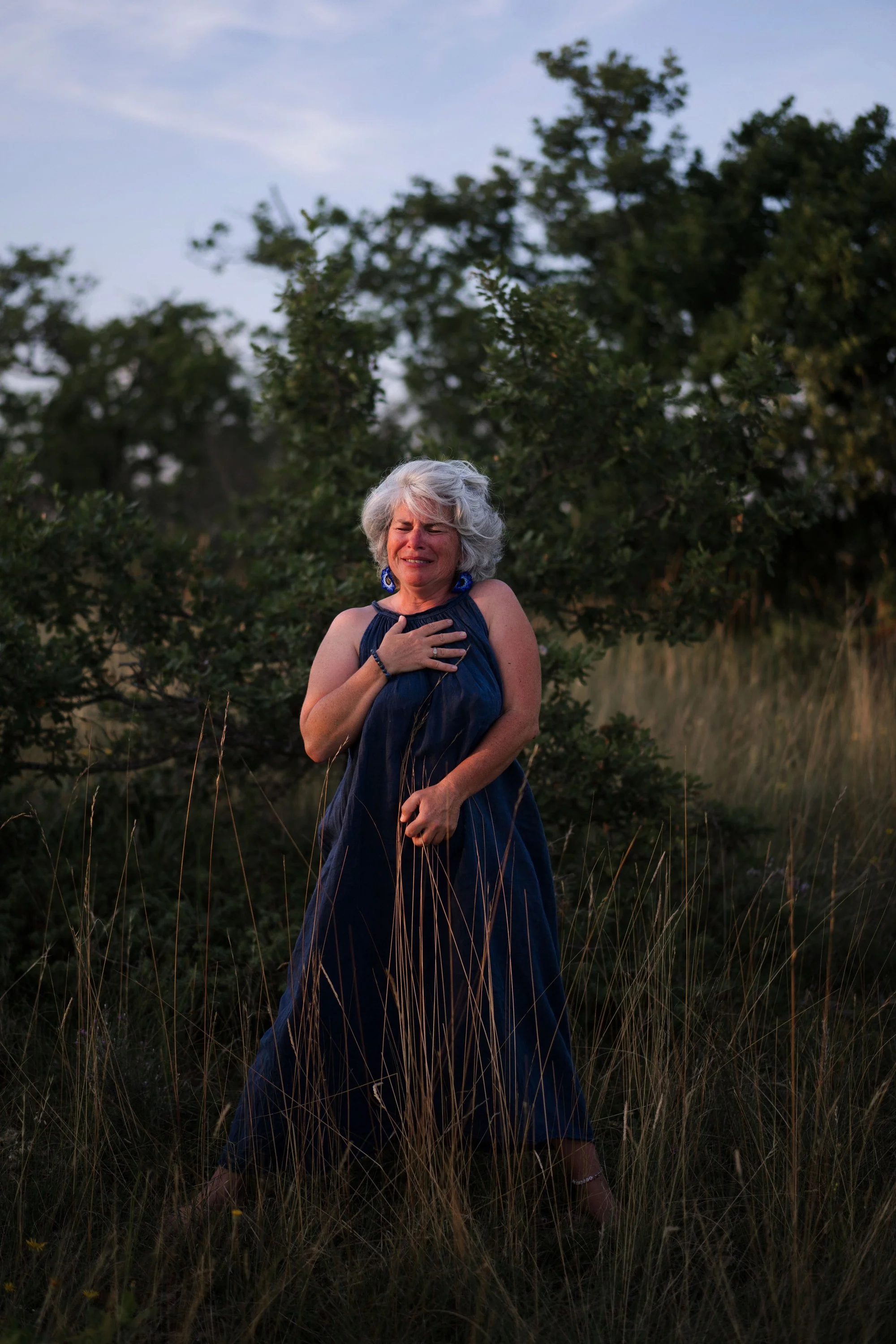 Une femme âgée portant une robe bleue, avec des cheveux gris, semble pleurer ou être émue, tandis qu'elle se tient dans une prairie entourée d'arbres, au crépuscule ou au coucher du soleil.