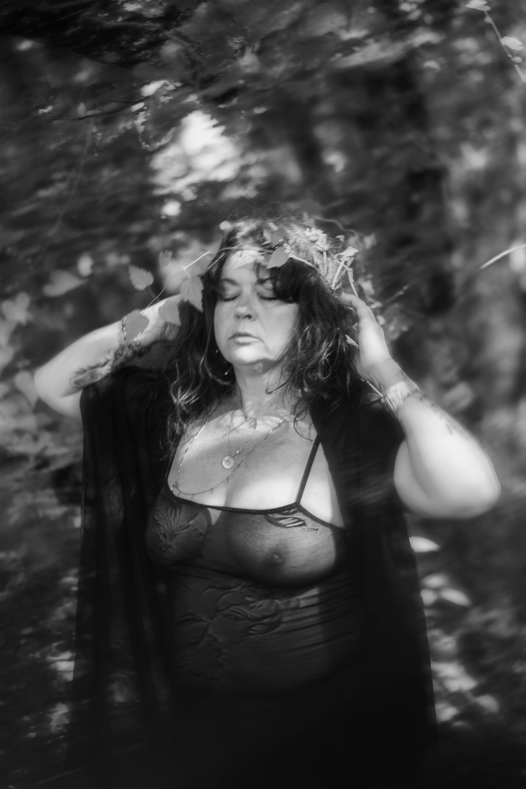 Une femme avec des cheveux longs et foncés, portant un haut en dentelle et un kimono ou un vêtement léger, pose devant un fond flou de feuilles d'arbres, avec un air sérieux et les yeux fermés, créé en mode monochrome.