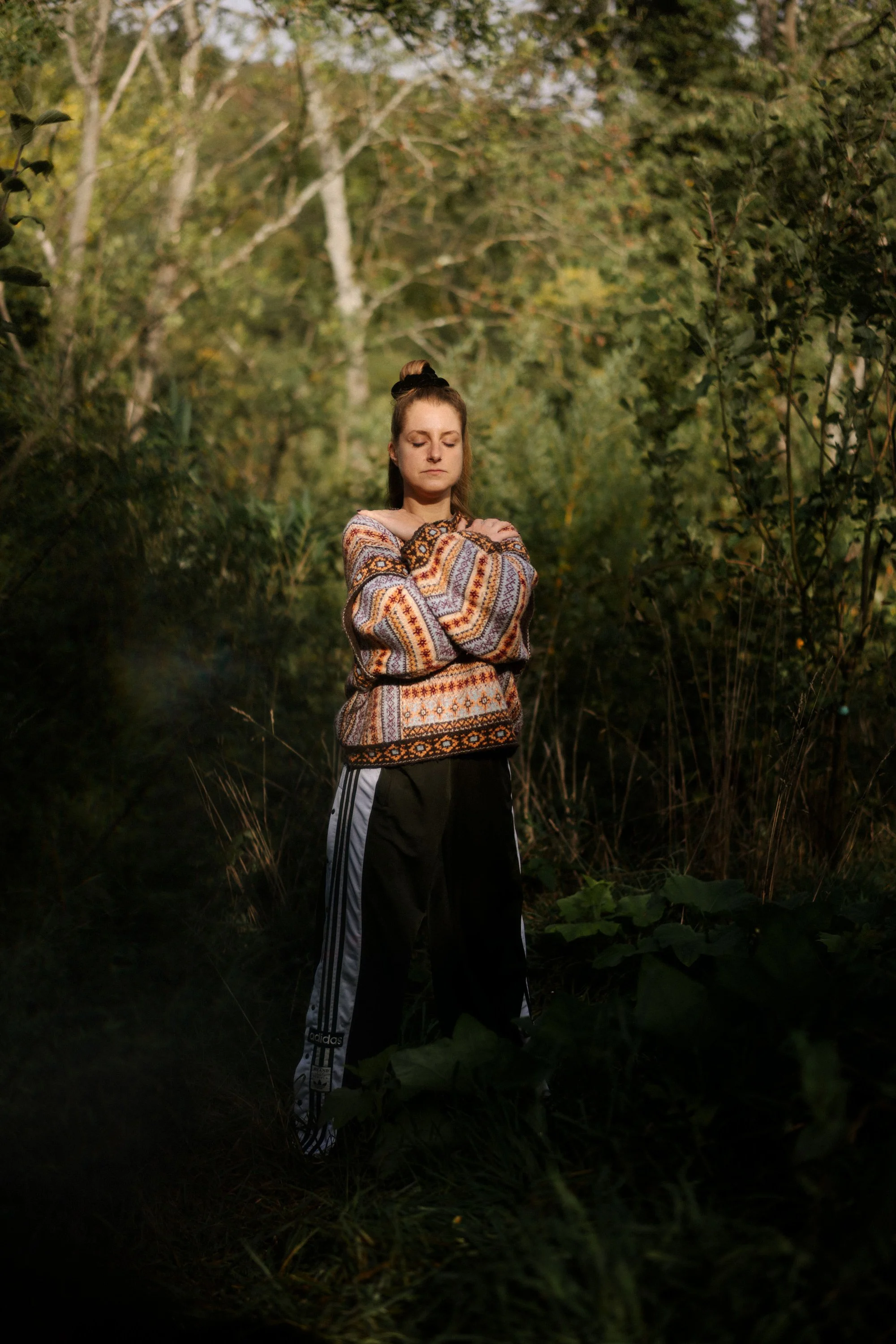Une jeune femme avec les bras croisés, portant un sweat coloré et un pantalon de sport, se tient dans une forêt en automne, le regard fermé, avec des arbres aux feuilles jaunes et vertes derrière elle.