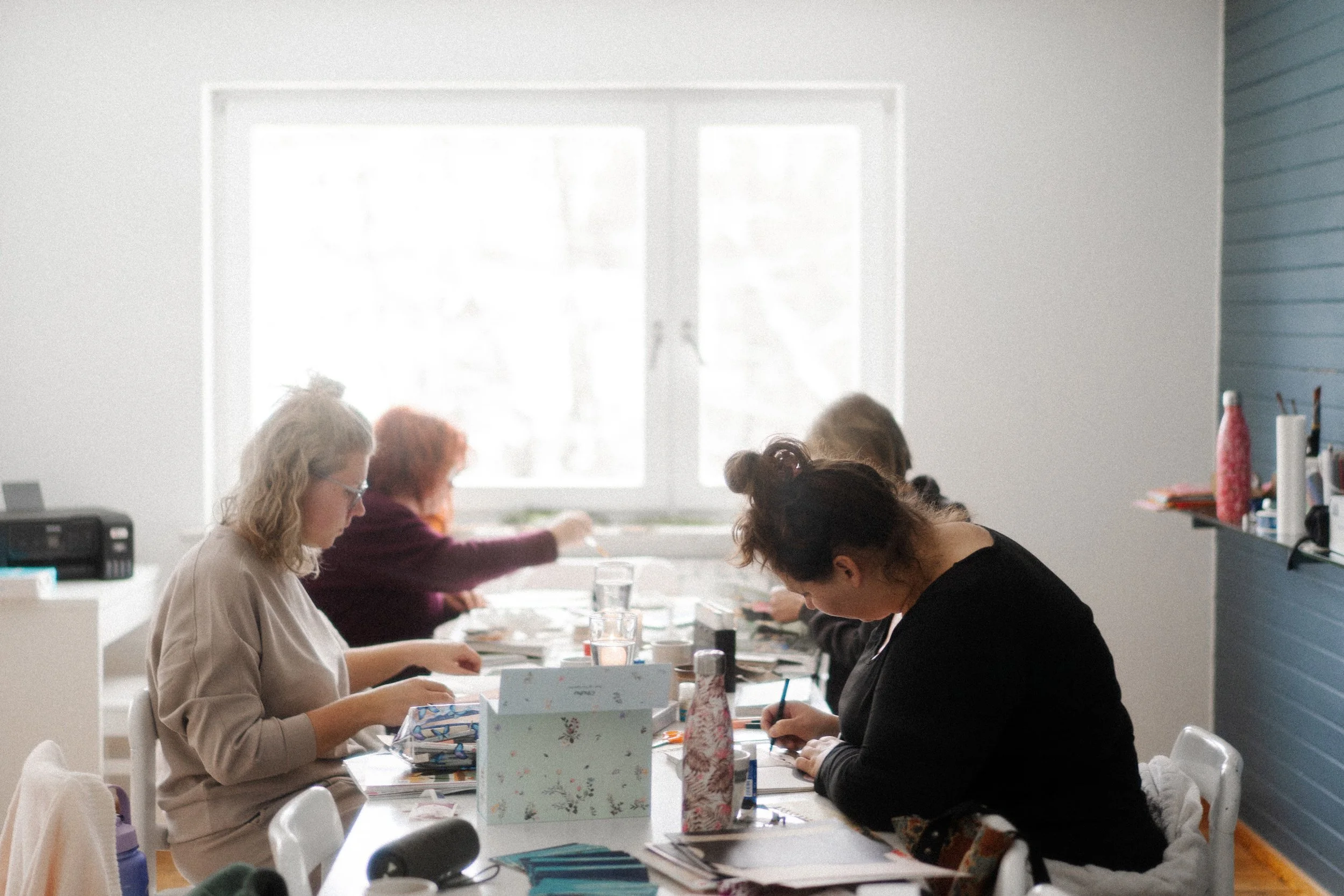 Groupe de personnes assises autour d'une table, concentrées sur une activité artistique ou manuelle dans une pièce lumineuse avec des fenêtres blanches.