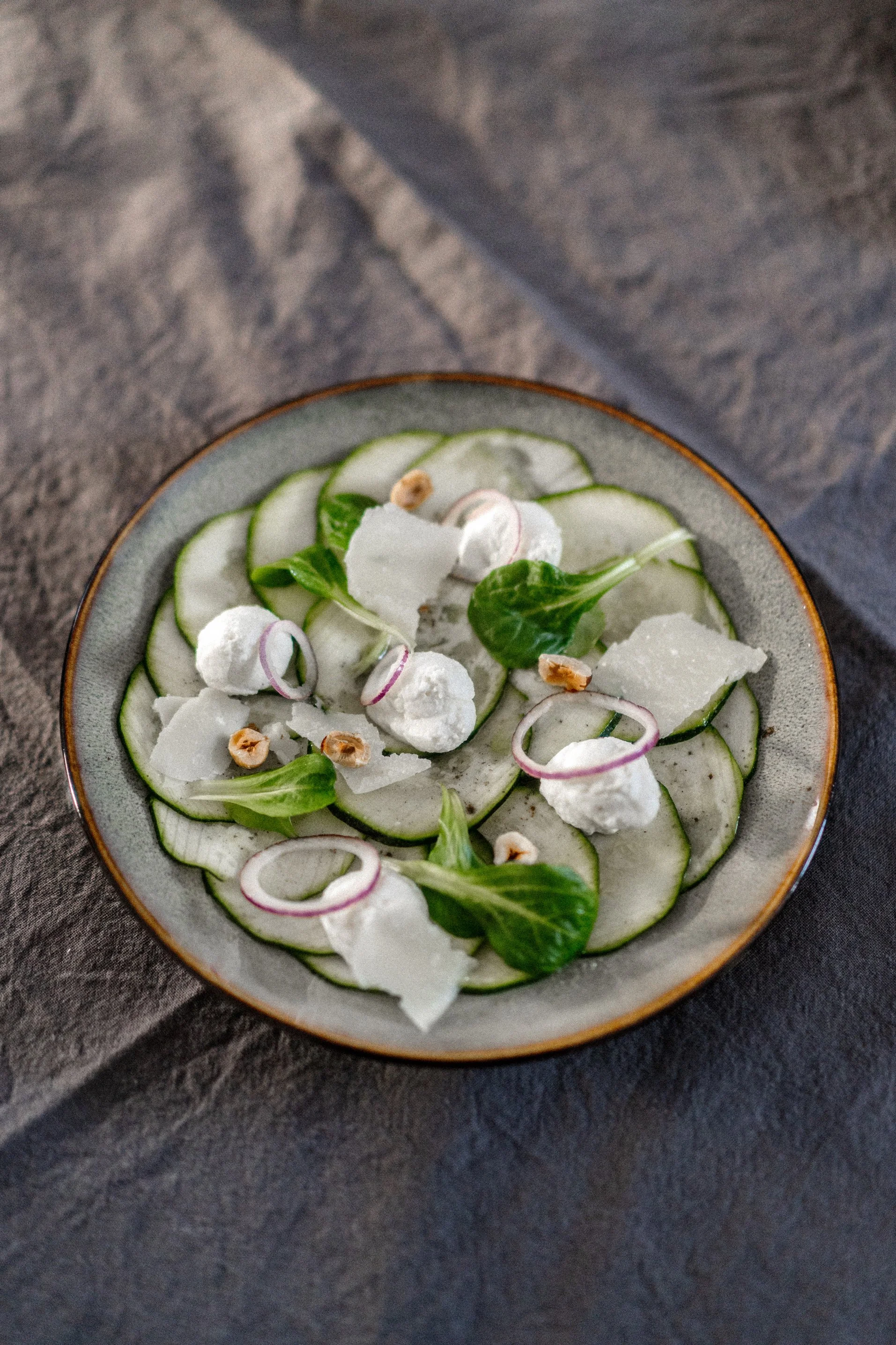 Une salade de concombre, fromage, oignons rouges, et noix dans un bol en céramique, posé sur une surface sombre.