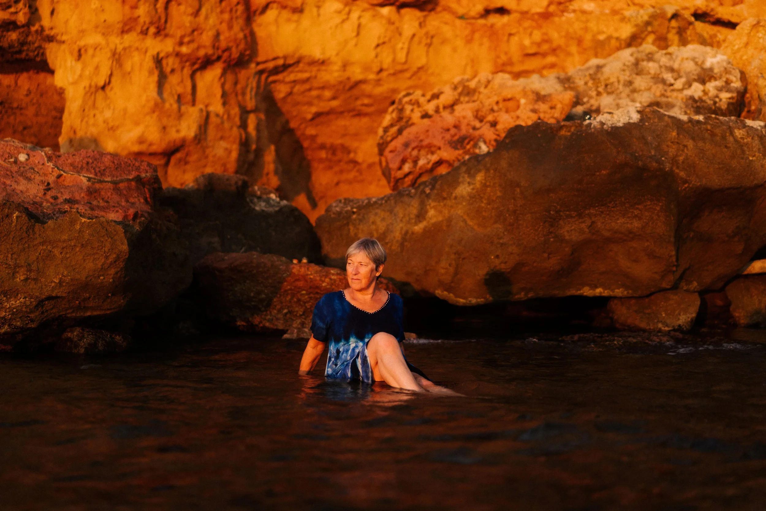 Une femme assise dans l'eau devant un fond rocheux orange et rouge, vraisemblablement dans une grotte ou un site naturel.
