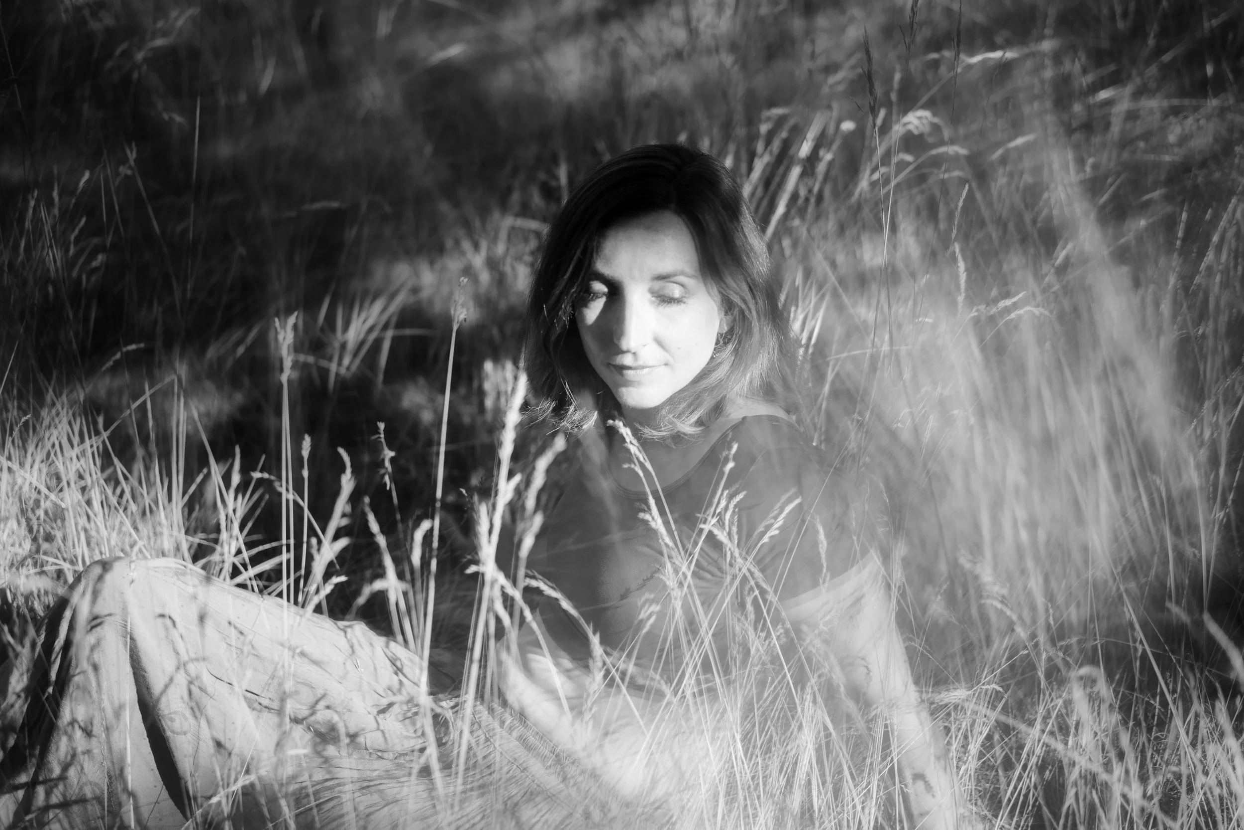 Une femme avec des cheveux bouclés, assise dans une prairie sèche. L'image est en noir et blanc, avec la femme au centre entourée de hautes herbes et de branches.