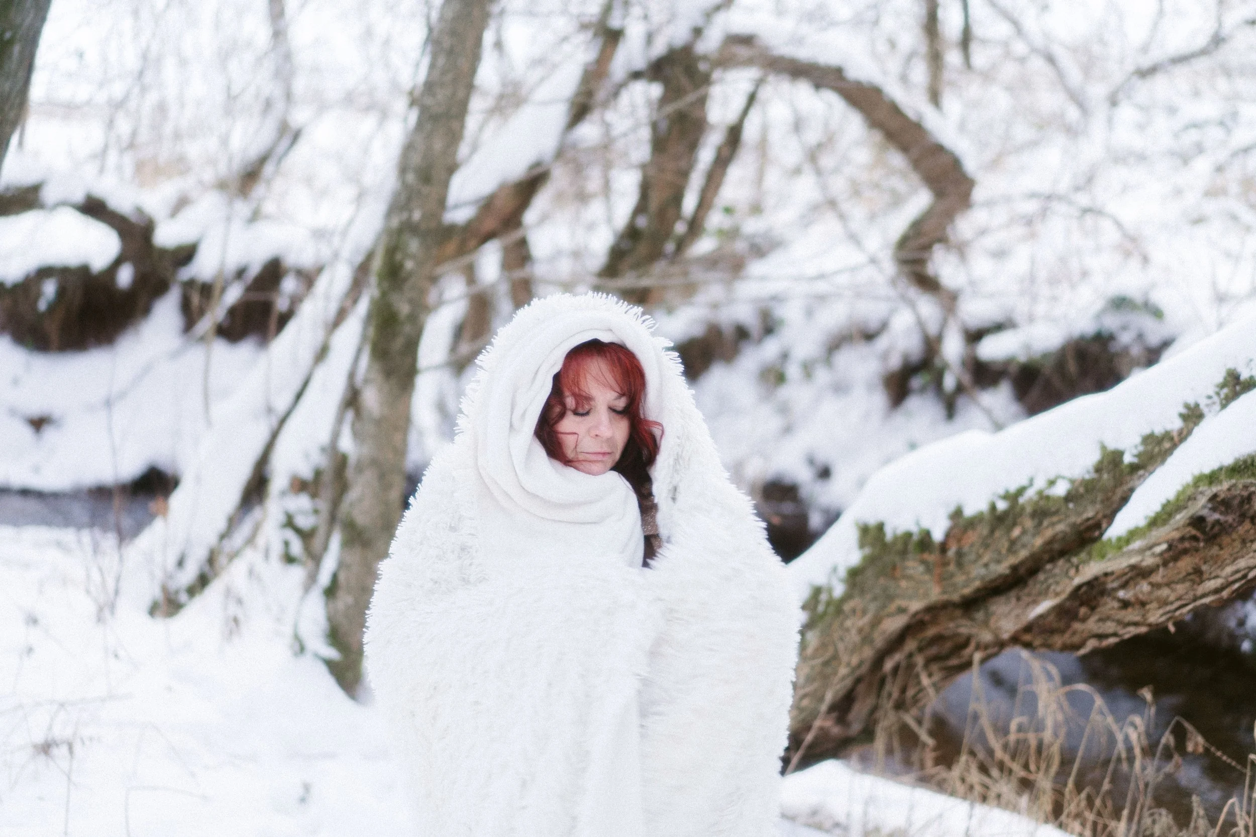 Femme avec un manteau blanc et une capuche en laine, dans un paysage enneigé avec des arbres. Elle est vue de face avec les yeux fermés.