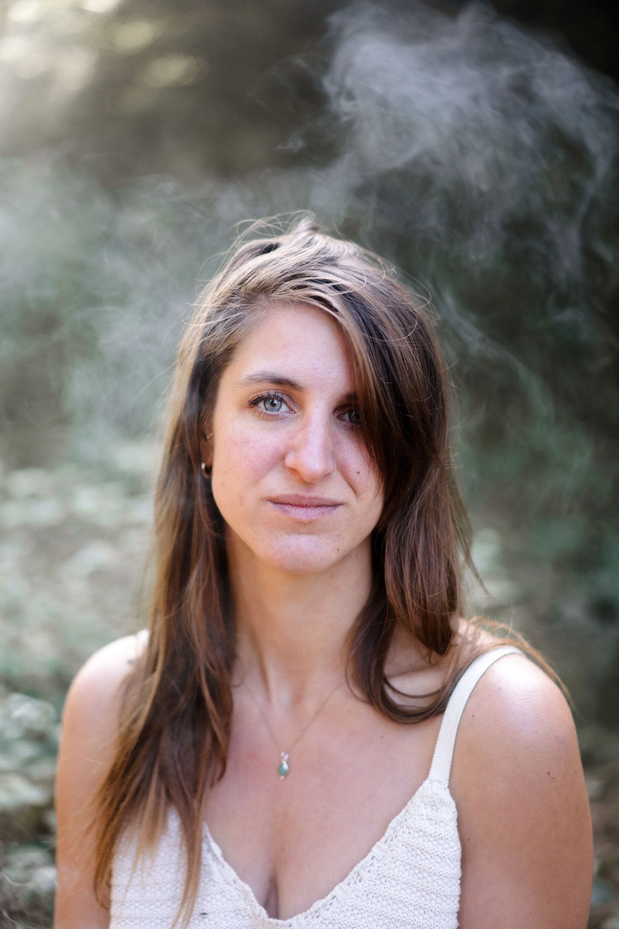 jeune femme aux cheveux longs et bruns, portant un haut blanc, en extérieur, avec un fond flou naturel.
