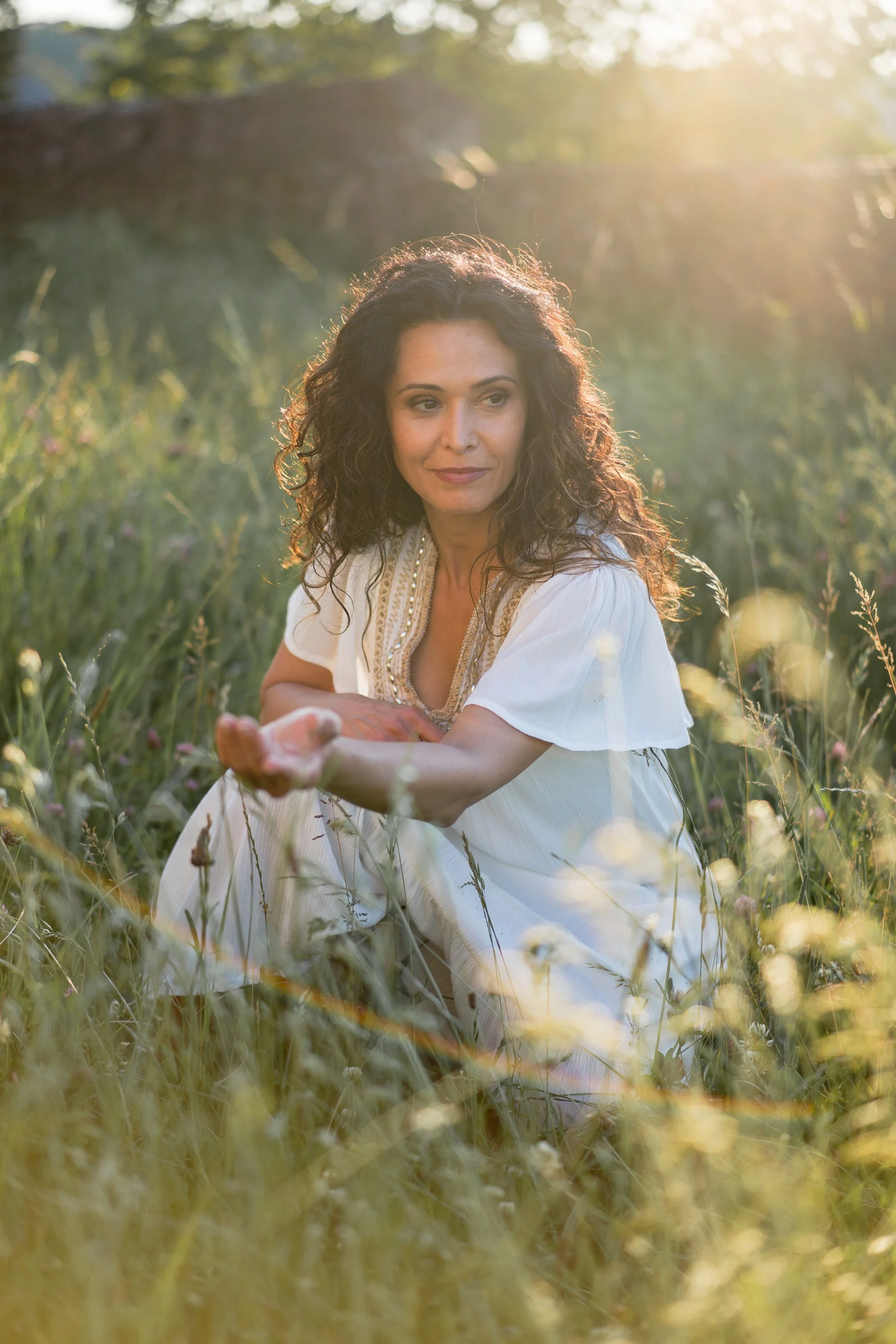 Une femme assise dans une prairie ensoleillée, portant une robe blanche, entourée de fleurs et d'herbes hautes.
