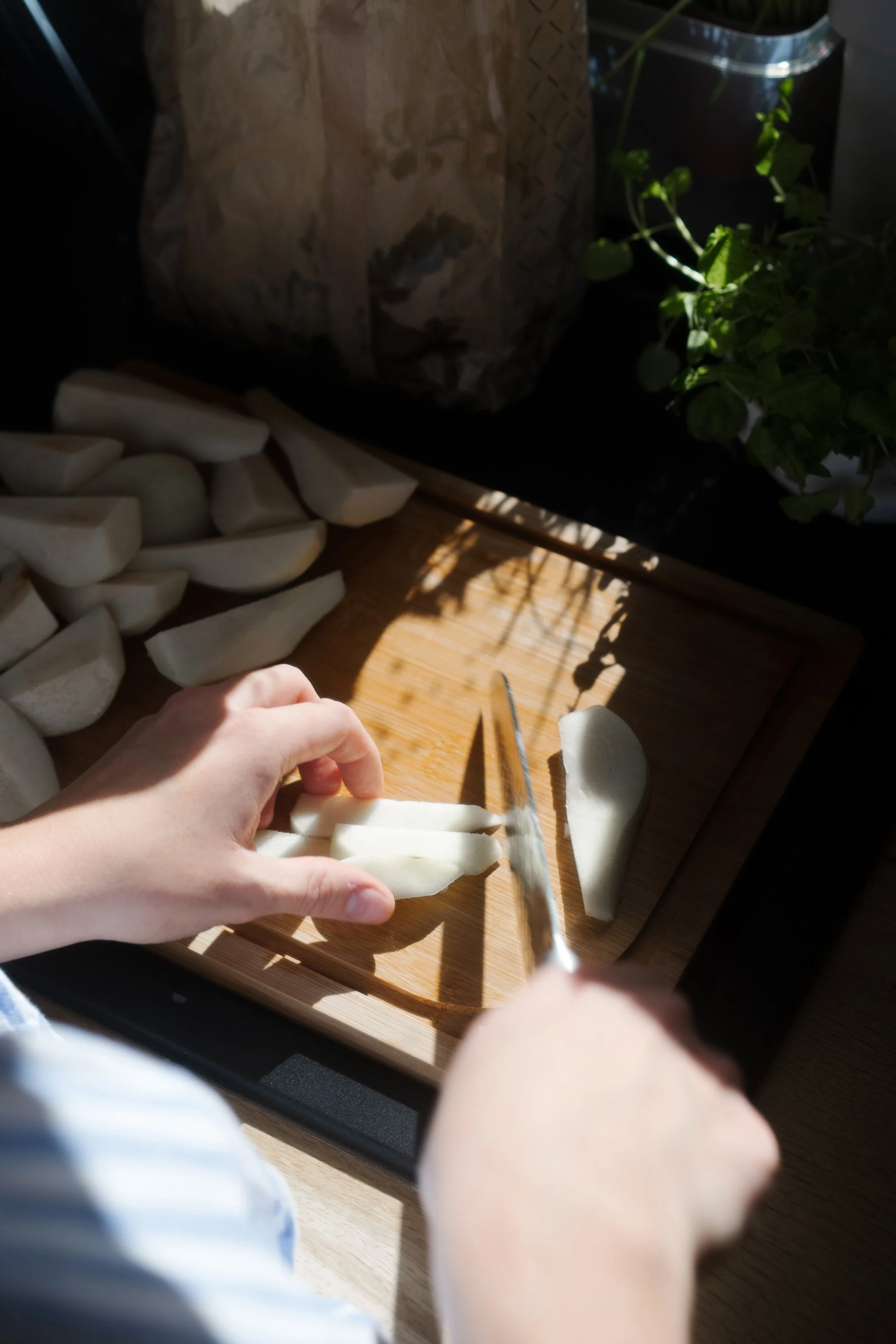 Une personne coupe des légumes blancs, probablement des navets, sur une planche en bois. Il y a d'autres légumes déjà coupés à côté et un plat avec des restes de légumes. La scène se déroule près d'une fenêtre avec des plantes vertes à l'arrière-plan.