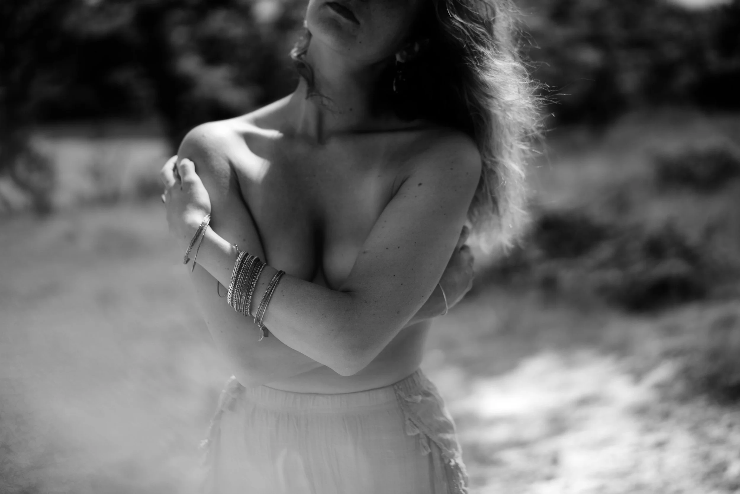Image en noir et blanc d'une femme qui se tient dans la nature, les bras croisés sur la poitrine, cheveux ondulés et portant plusieurs bracelets, avec un visage légèrement visible et une gorge nue.