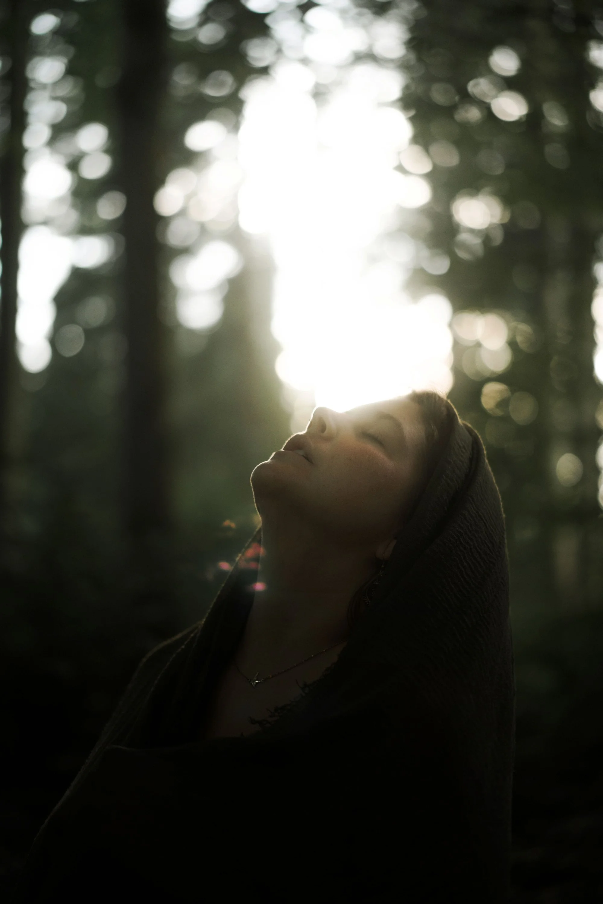 Une personne avec un hoodie, yeux fermés, se tenant dans une forêt avec la lumière du soleil qui filtre à travers les arbres.