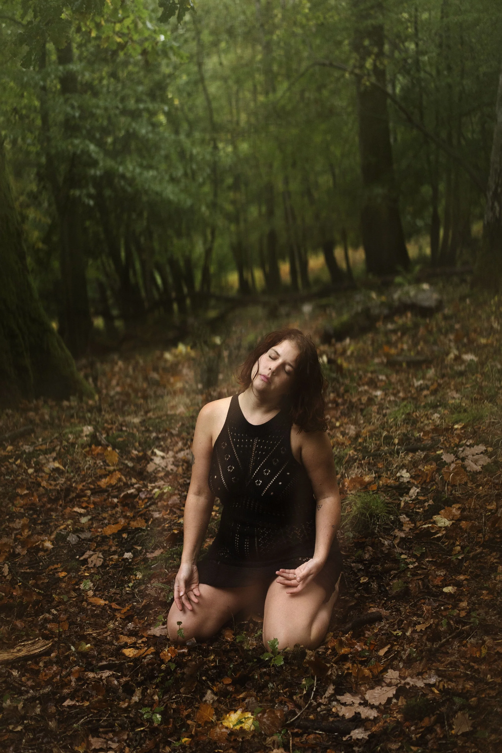 Une femme assise sur le sol en forêt, porte un pantalon noir et un haut noir, avec des feuilles mortes autour d'elle, arrière-plan dense d'arbres verts.