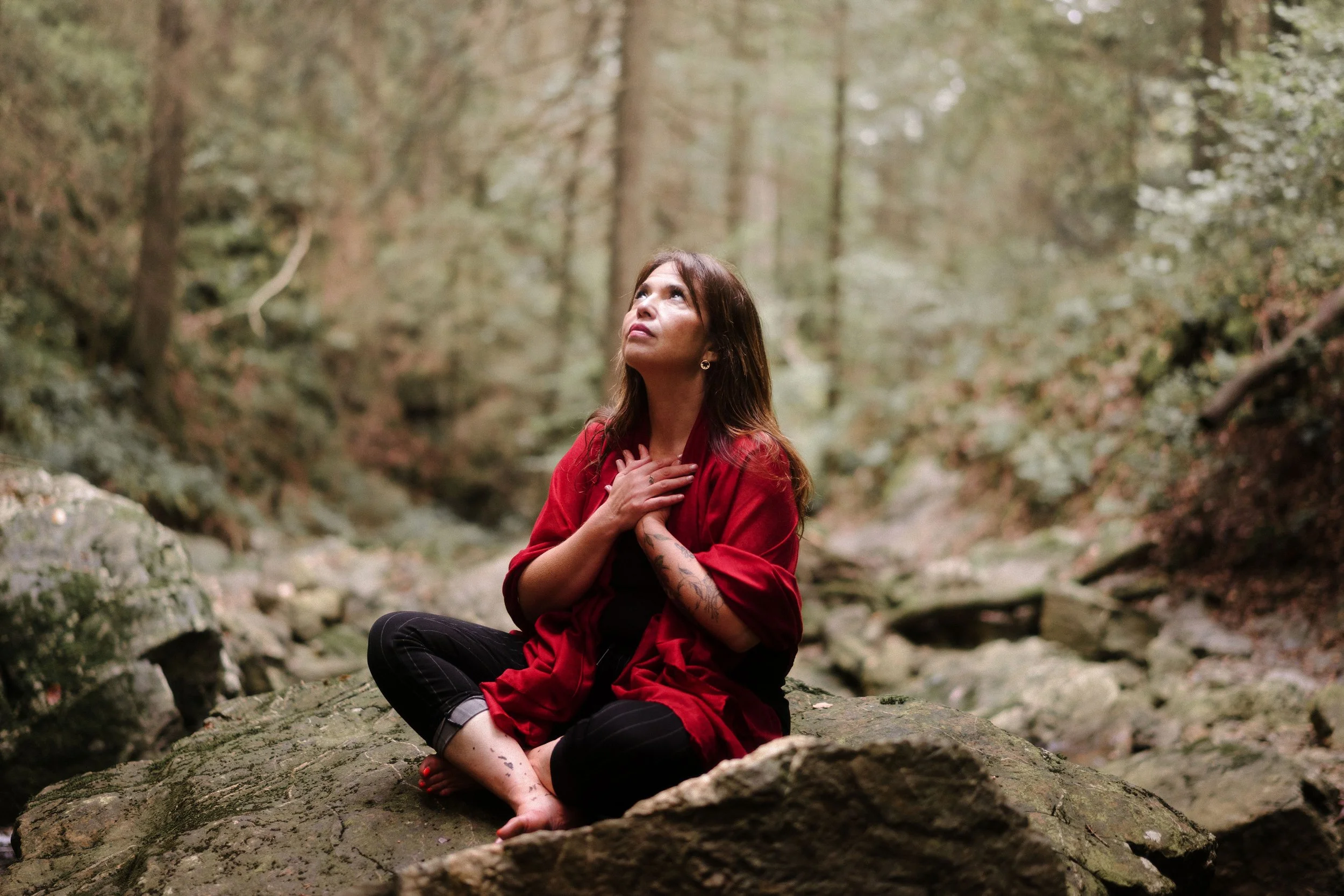 Femme assise sur une roche dans une forêt, portant un pull rouge, regardant vers le haut, mains sur la poitrine.