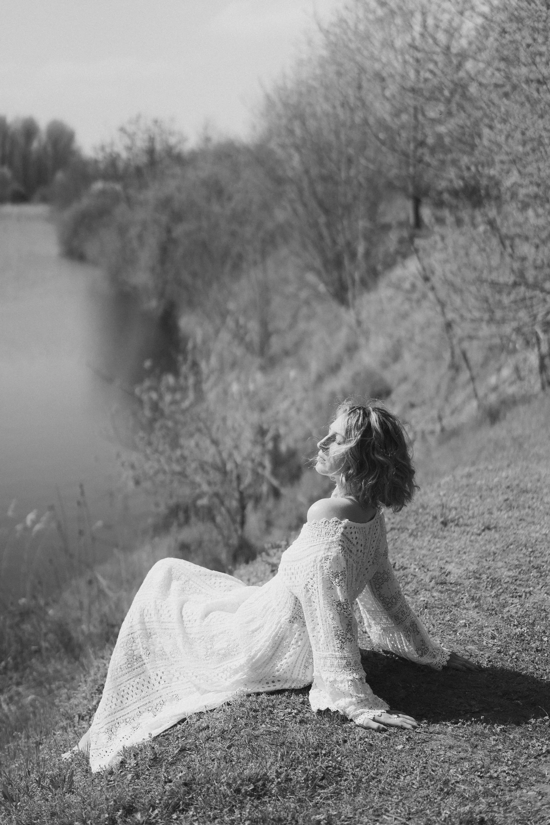 Femme assise sur l'herbe dans un champ, portant une robe longue en dentelle blanche, avec des arbres en arrière-plan, photographiée en noir et blanc.